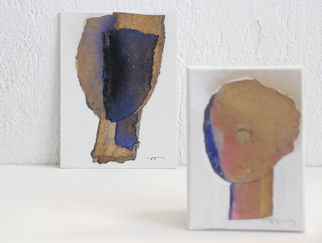 Two abstract watercolor paintings, one on canvas on the left with a central dark blue shape, and one on a small rectangular canvas on the right depicting a stylized portrait with a rounded head and a simple neck, both on a white surface with a textur