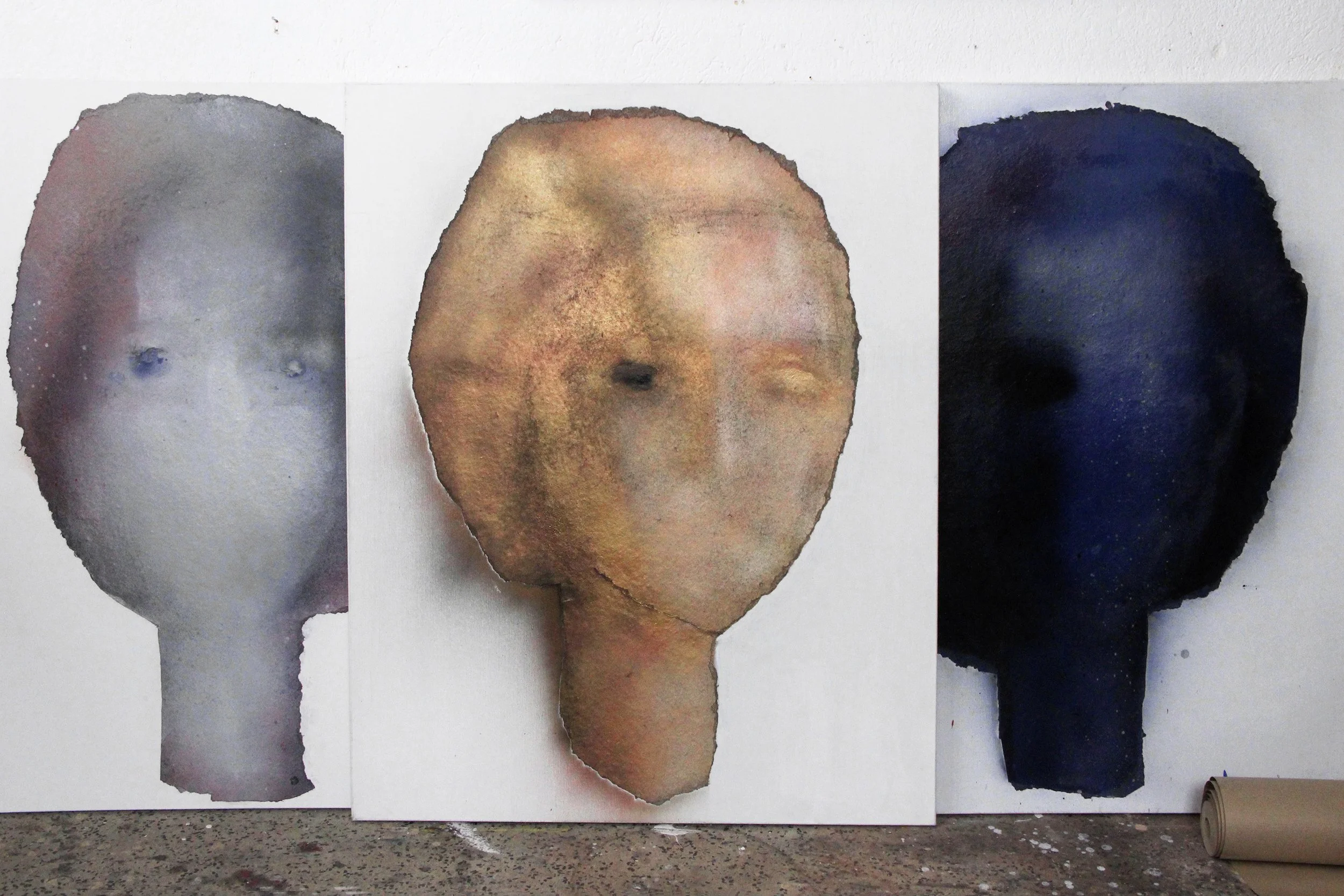 Three abstract watercolor paintings of human faces on white paper, each with distinct color schemes, arranged side by side on a wall.