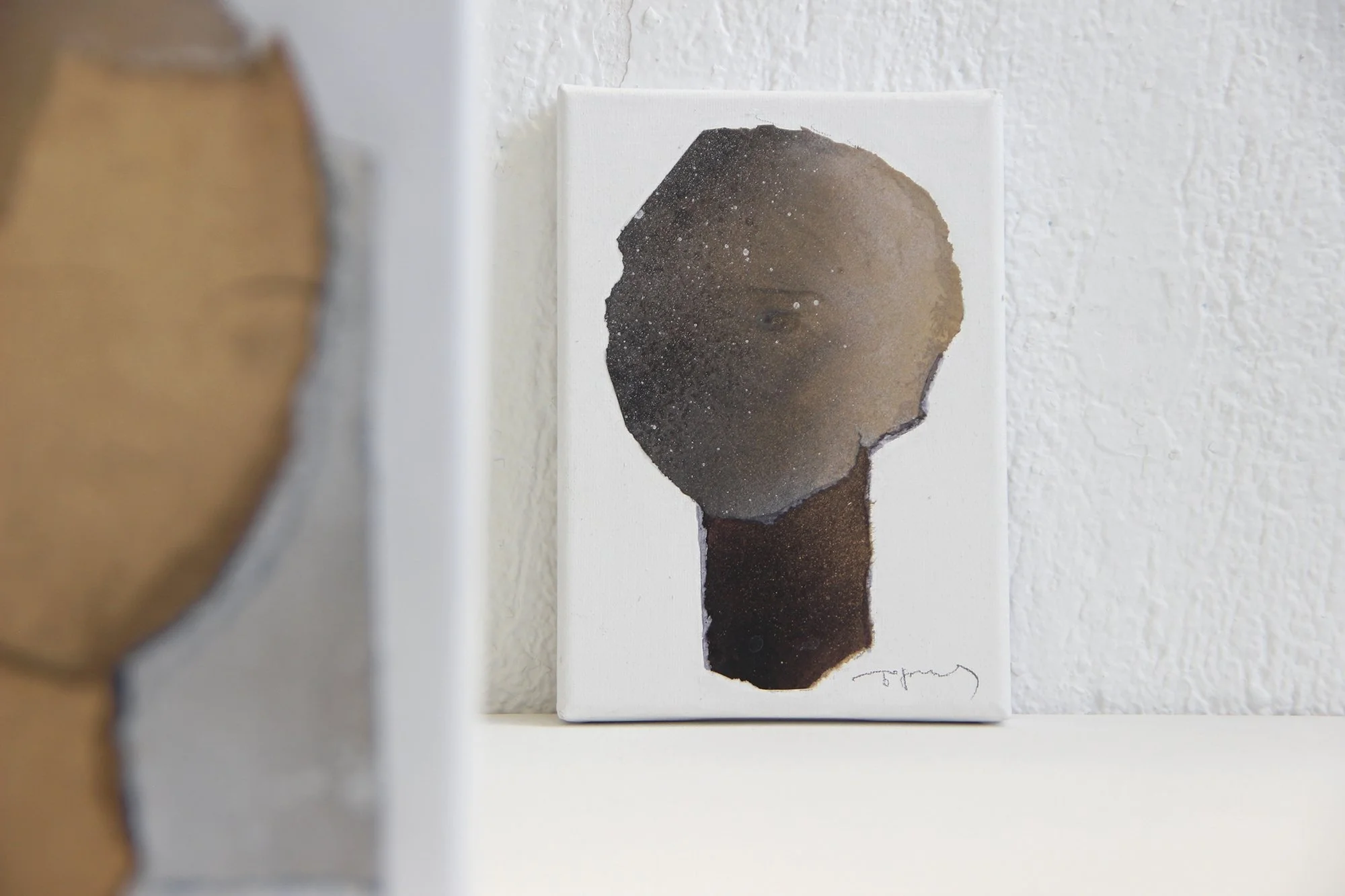 Small abstract painting of a human head with a dark face and textured brown neck, signed at the bottom right, leaning against a textured white wall.