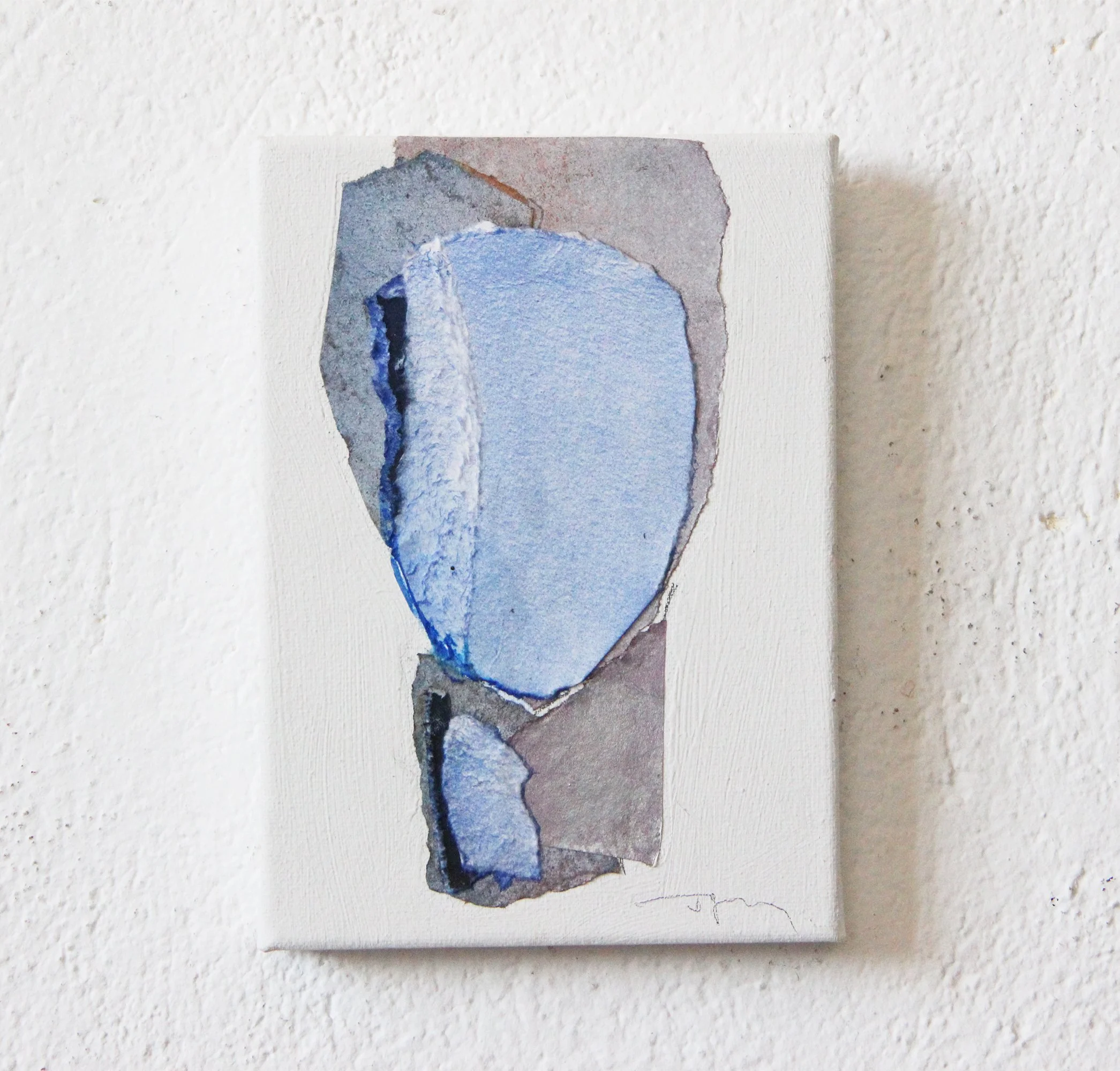 Abstract watercolor painting of blue and gray shapes on a white canvas, hanging on a textured white wall.