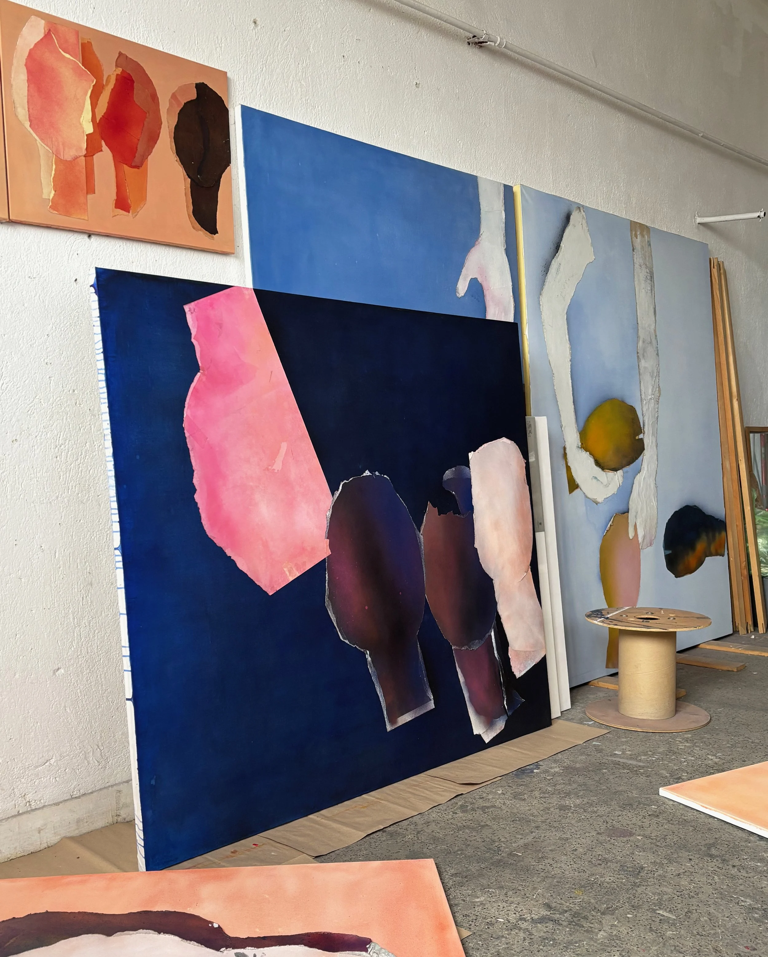 Multiple abstract paintings displayed in a studio, featuring torn paper-like textures and organic shapes in vibrant colors such as pink, black, white, blue, and earth tones.