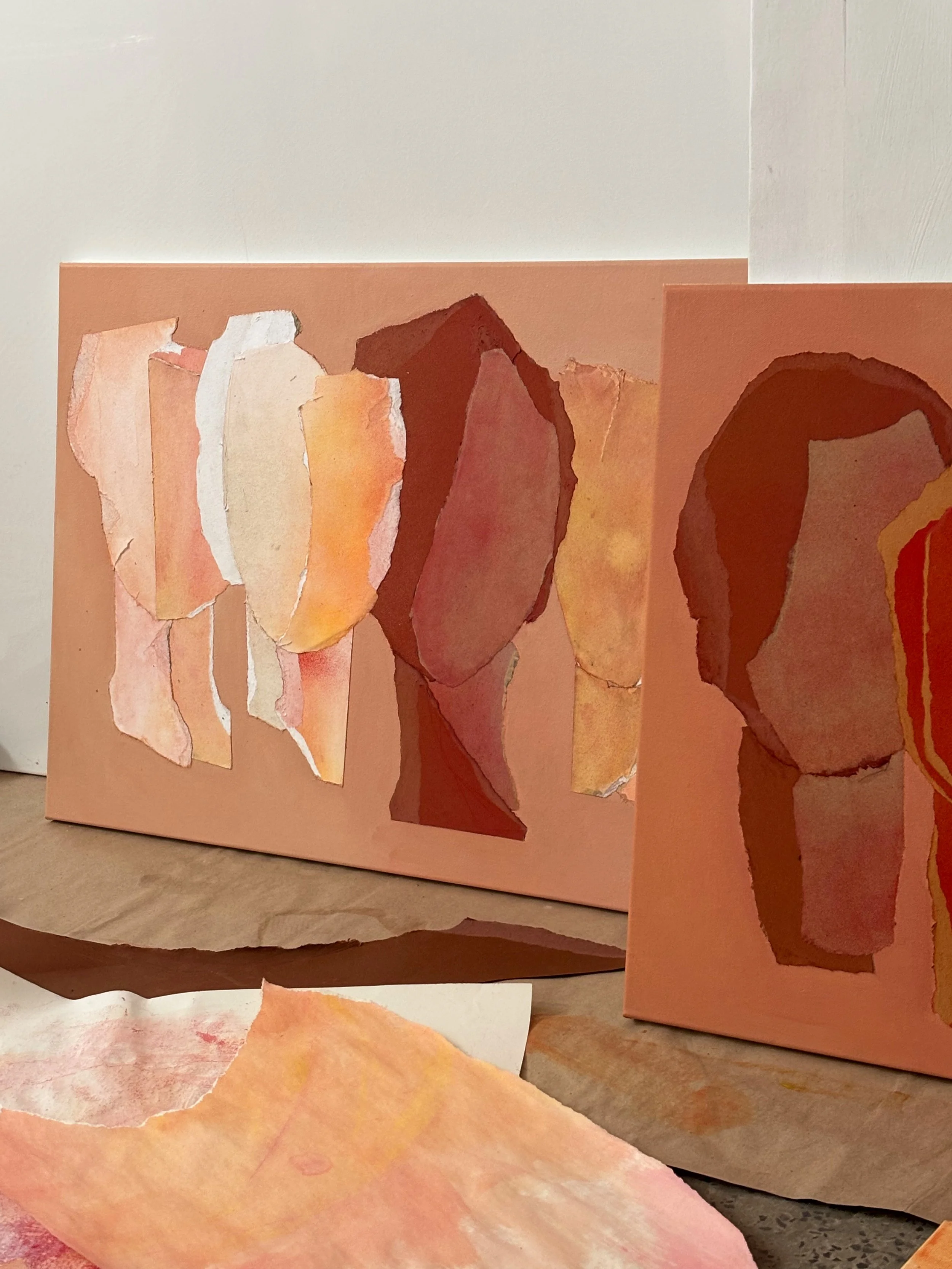 Two abstract watercolor paintings on paper, featuring warm tones of pink, peach, orange, brown, and red, with torn edges and layered textures, leaning against a wall on a wooden surface.