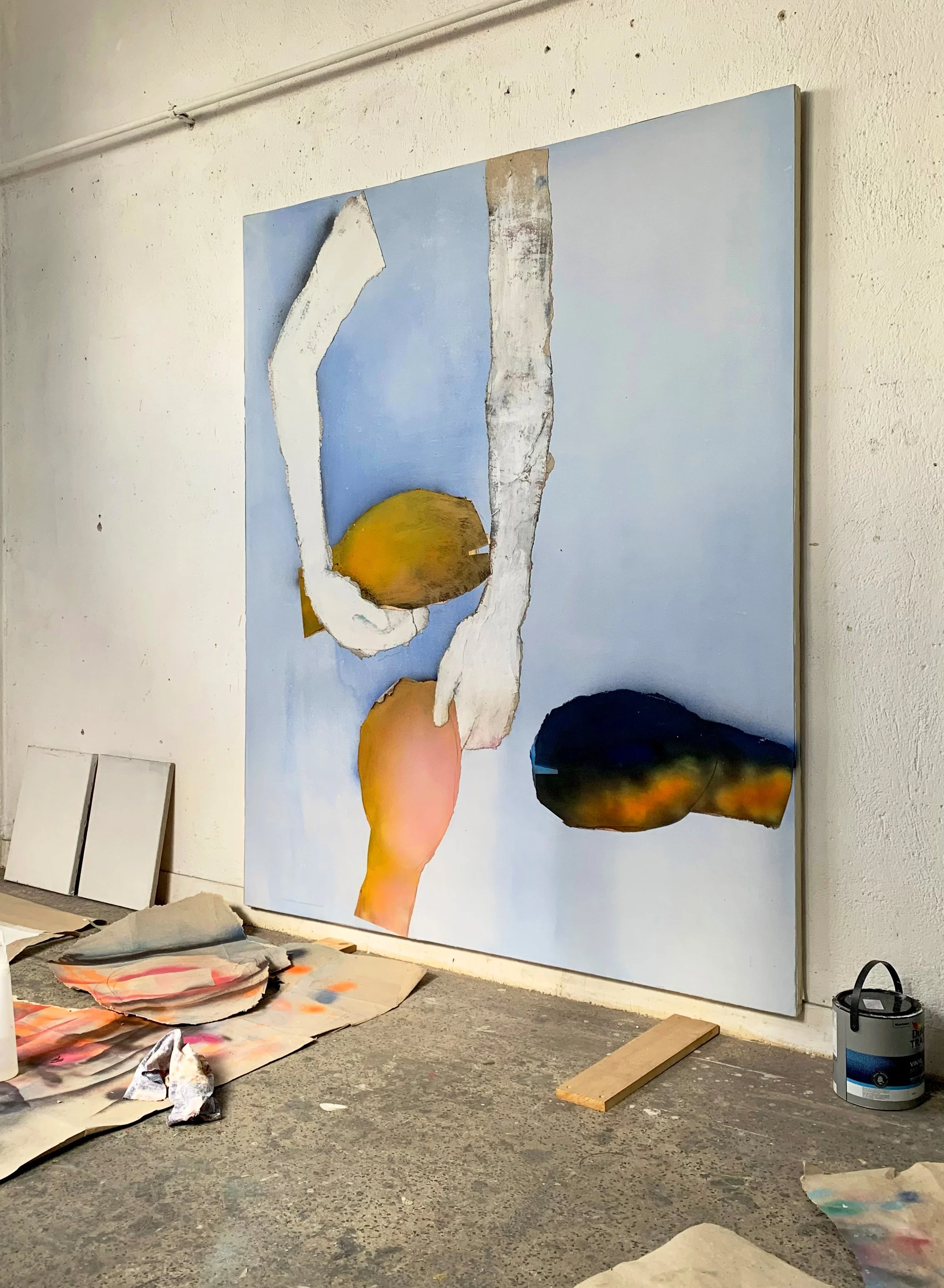 An abstract painting on a large canvas features disconnected shapes of color on a light blue background, with art supplies and canvases on the floor.