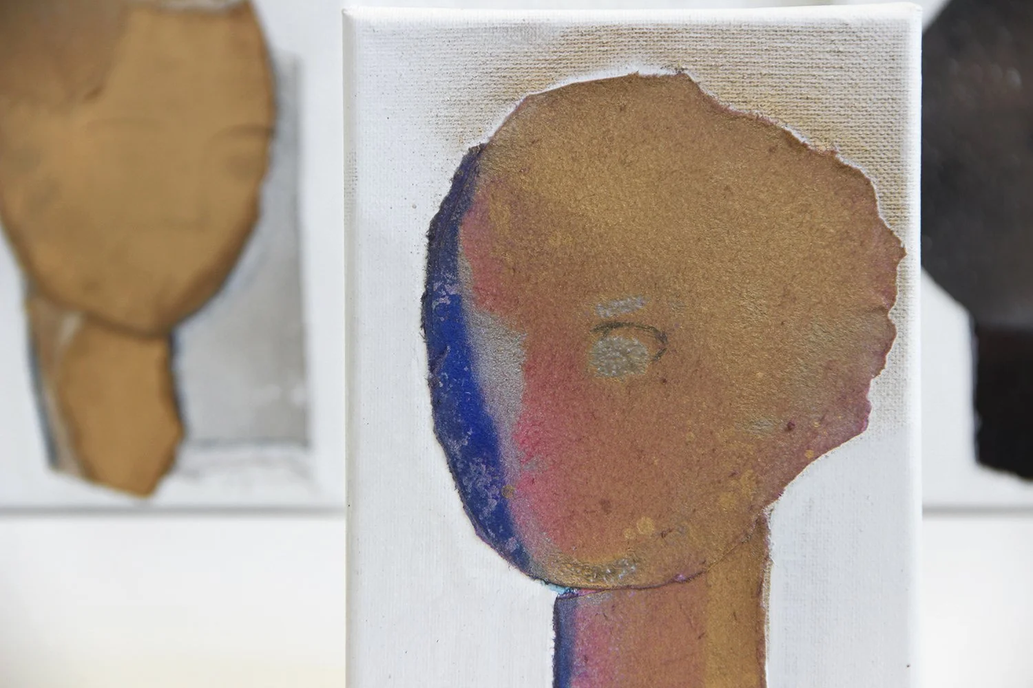 Abstract painting featuring a textured, circular shape in brown and red tones with hints of blue, on a white background, with two more blurred paintings in the background.