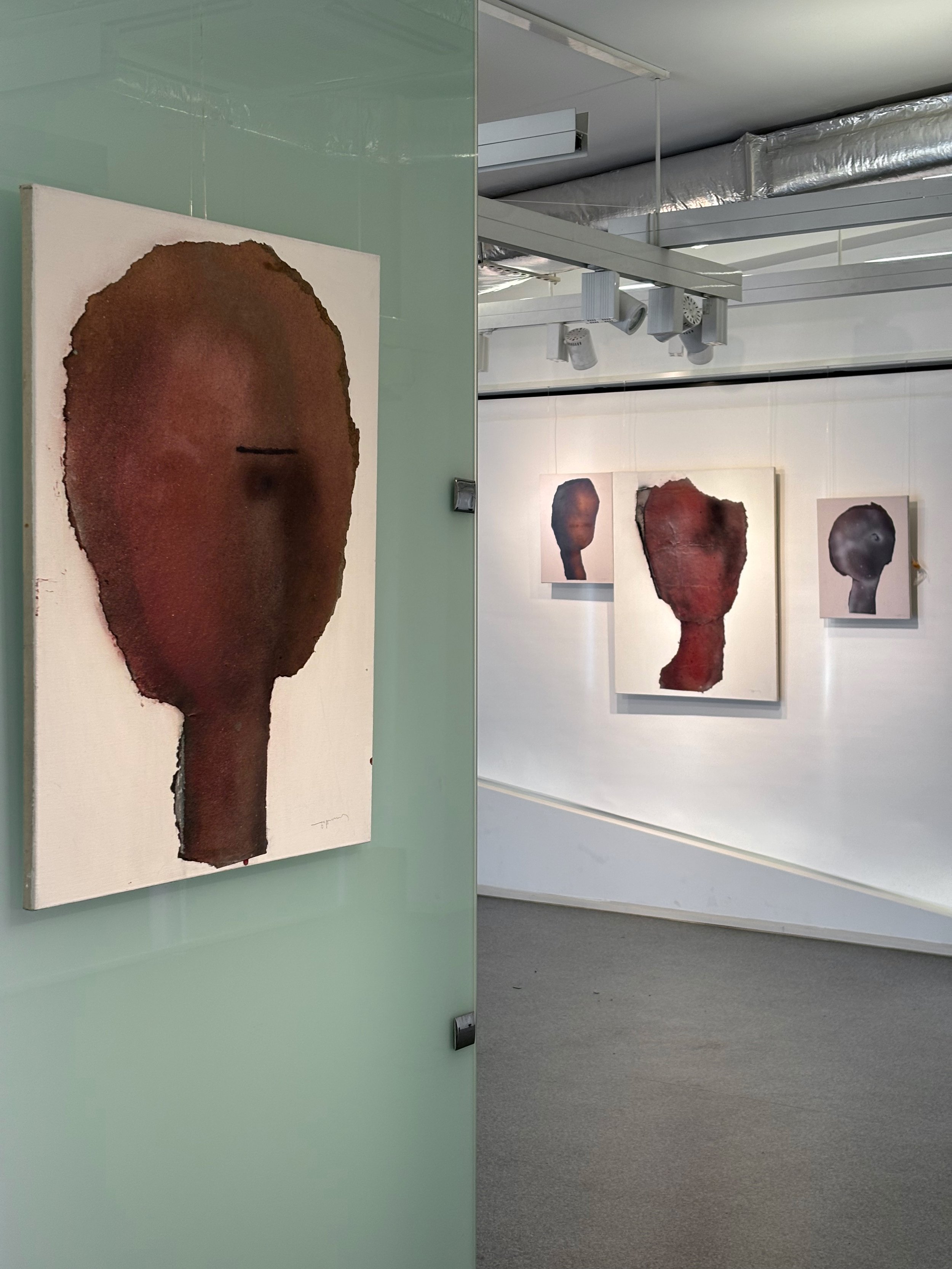 Art exhibit featuring abstract portrait paintings of human faces with textured, dark red and brown colors, displayed on white walls with ceiling tracks and lighting.