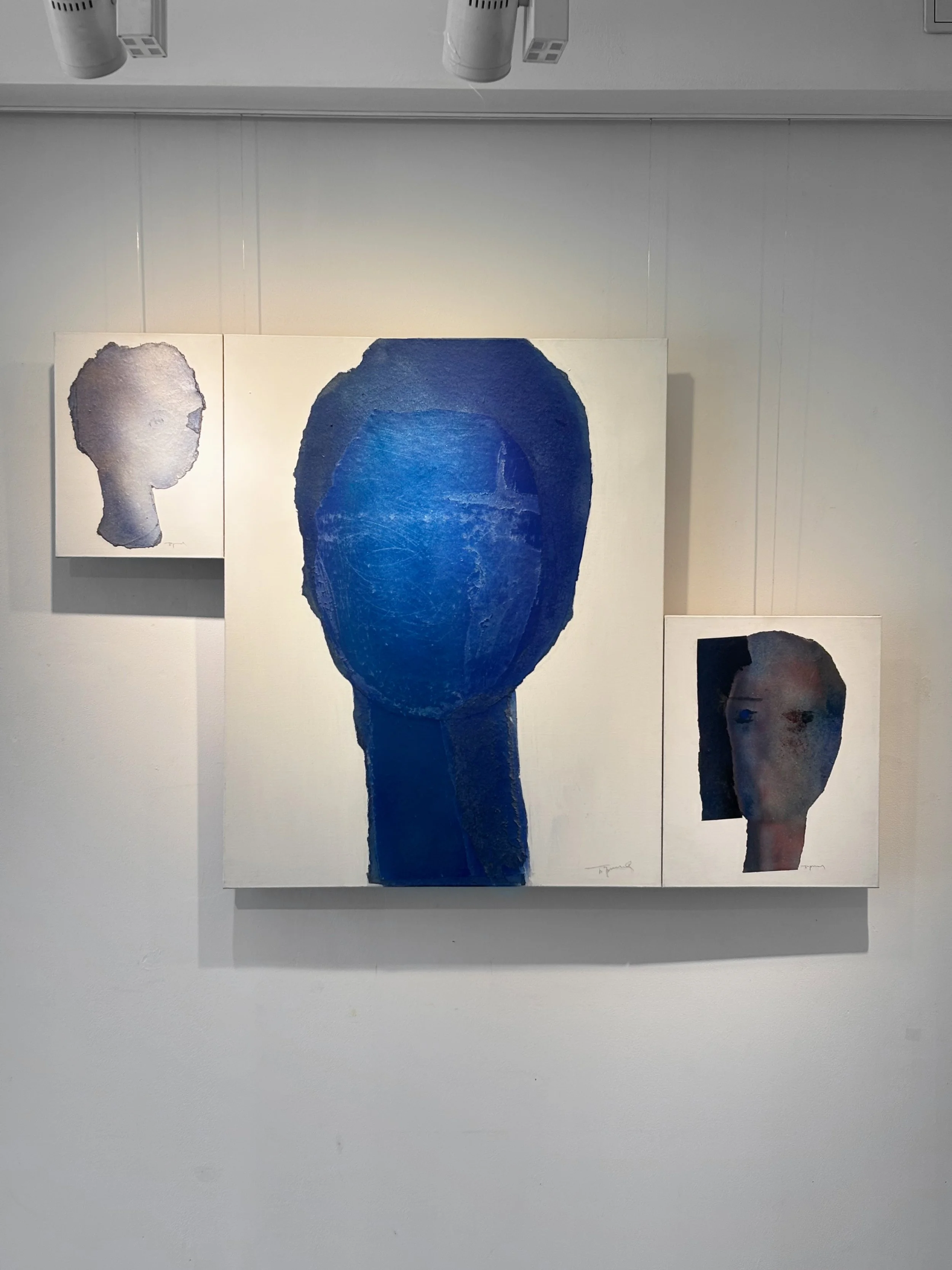 Three portrait paintings of human heads in an art gallery, with the central piece being a large blue figure without detailed facial features.