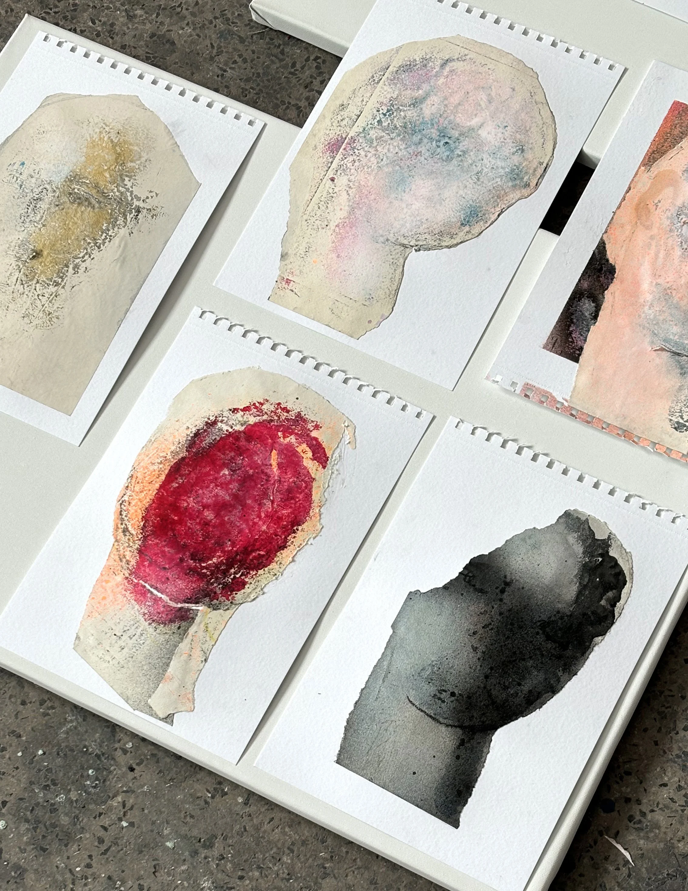 Collection of five abstract watercolor paintings of lightbulb silhouettes on white paper. Each features different color schemes: gold and gray, pastel pink and blue, red and peach, black and gray. They are laid out on a white surface.