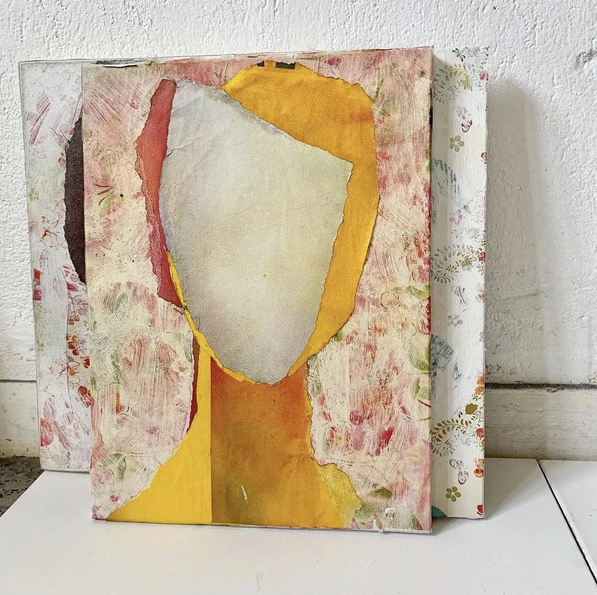 Abstract mixed media artwork with torn paper and paint in shades of yellow, pink, red, gray, and black, mounted on a textured wall.