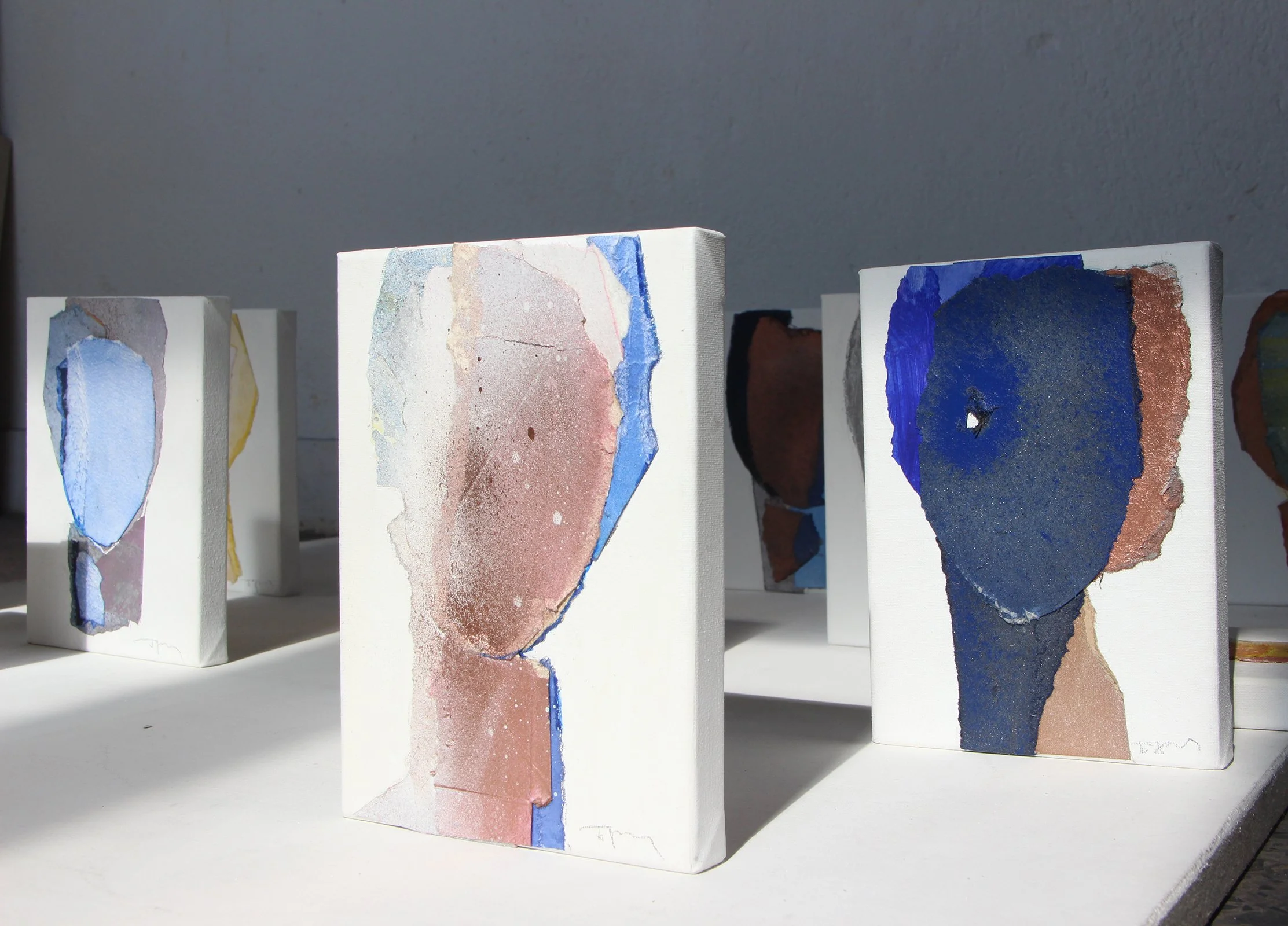 Small abstract paintings on canvas depicting minimalist human profiles, arranged on a white shelf in a gallery setting, with shadows cast on a white wall.