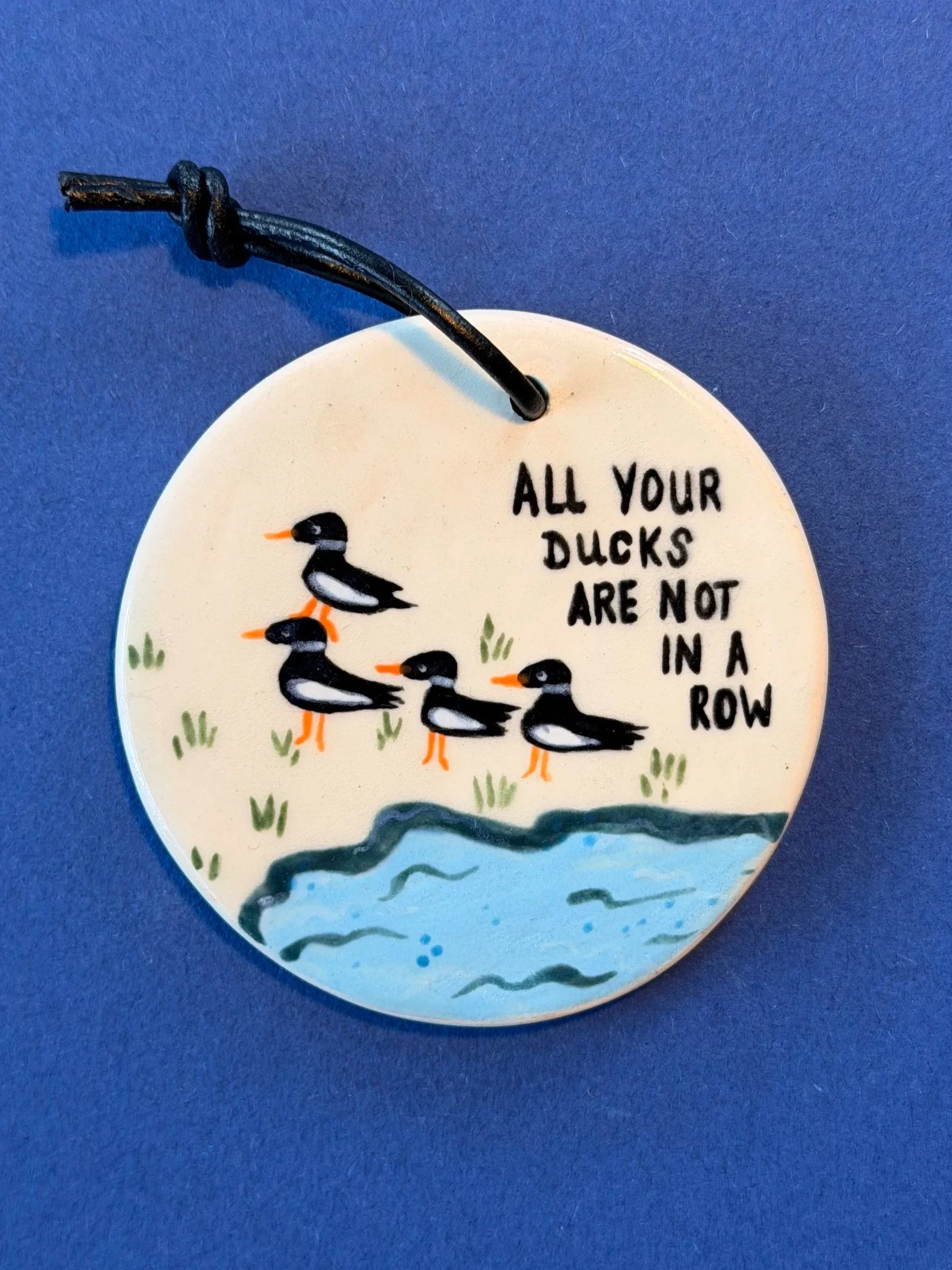 Decorative ceramic disk with a drawing of four ducks standing on grass near water, accompanied by the text 'All your ducks are not in a row', hanging on a blue background.