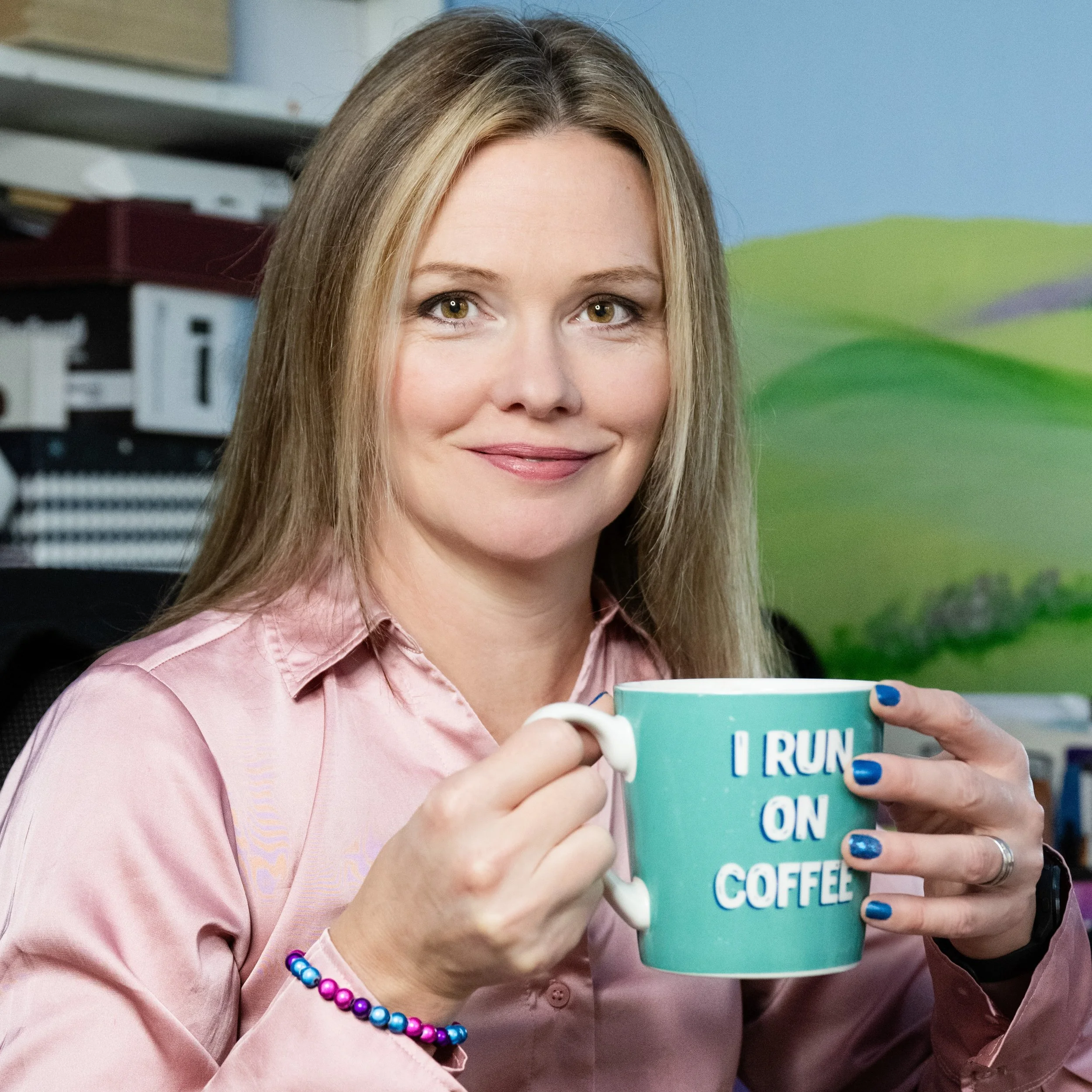 A woman with long blonde hair wearing a pink shirt and colorful beaded bracelet is holding a teal coffee mug with the text 'I run on coffee' printed on it. She has manicured nails painted blue and is smiling softly. The background suggests an office 
