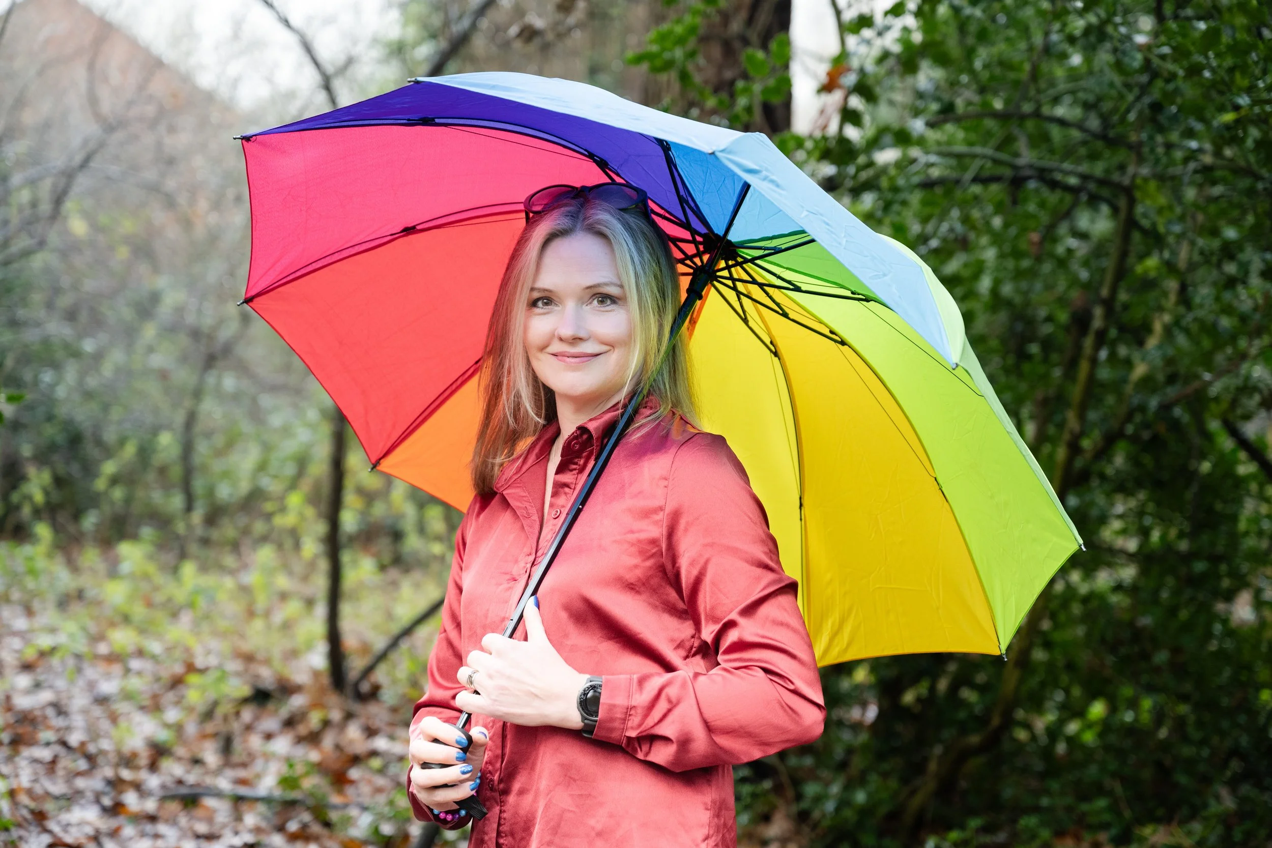 A woman smiling and holding a multicolored rainbow umbrella outdoors in a wooded area with fallen leaves on the ground.