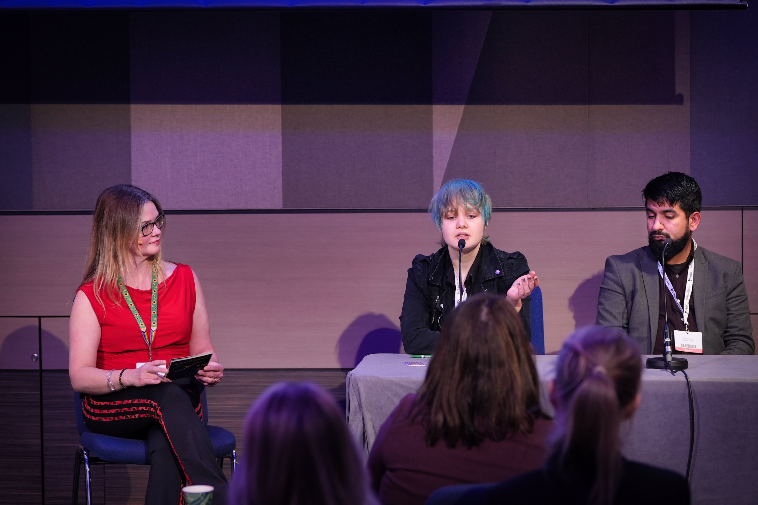 Three people sitting at a panel discussion, with an audience in front of them. The person in the middle has blue hair and is speaking into a microphone. The woman on the left has long hair and glasses, wearing a red top. The man on the right has dark
