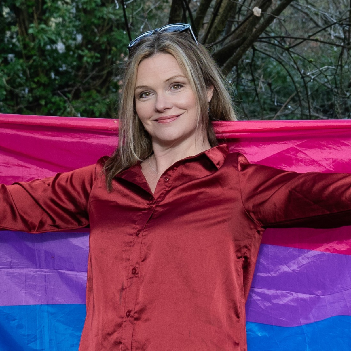 Charlie, a woman with shoulder-length dark blonde hair wearing a red satin shirt, holding a bi pride flag broad behind her with both arms extended.