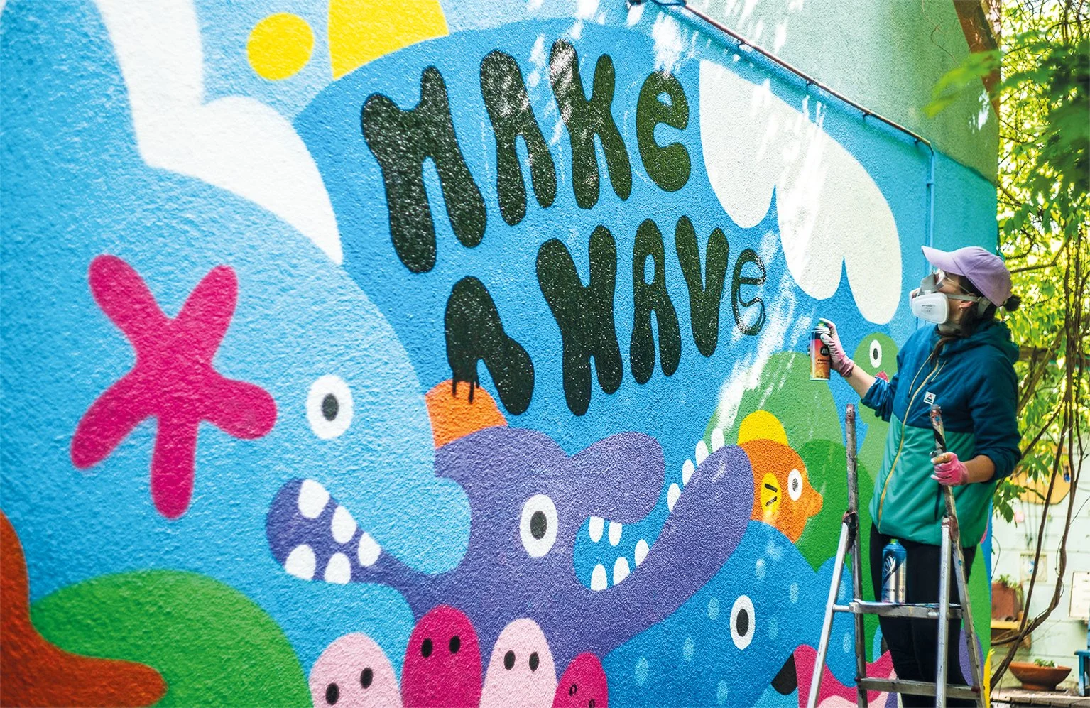 Make a wave - Wandbild. Künstlerin im Prozess besprüht eine Wand auf einer Leiter stehend.
