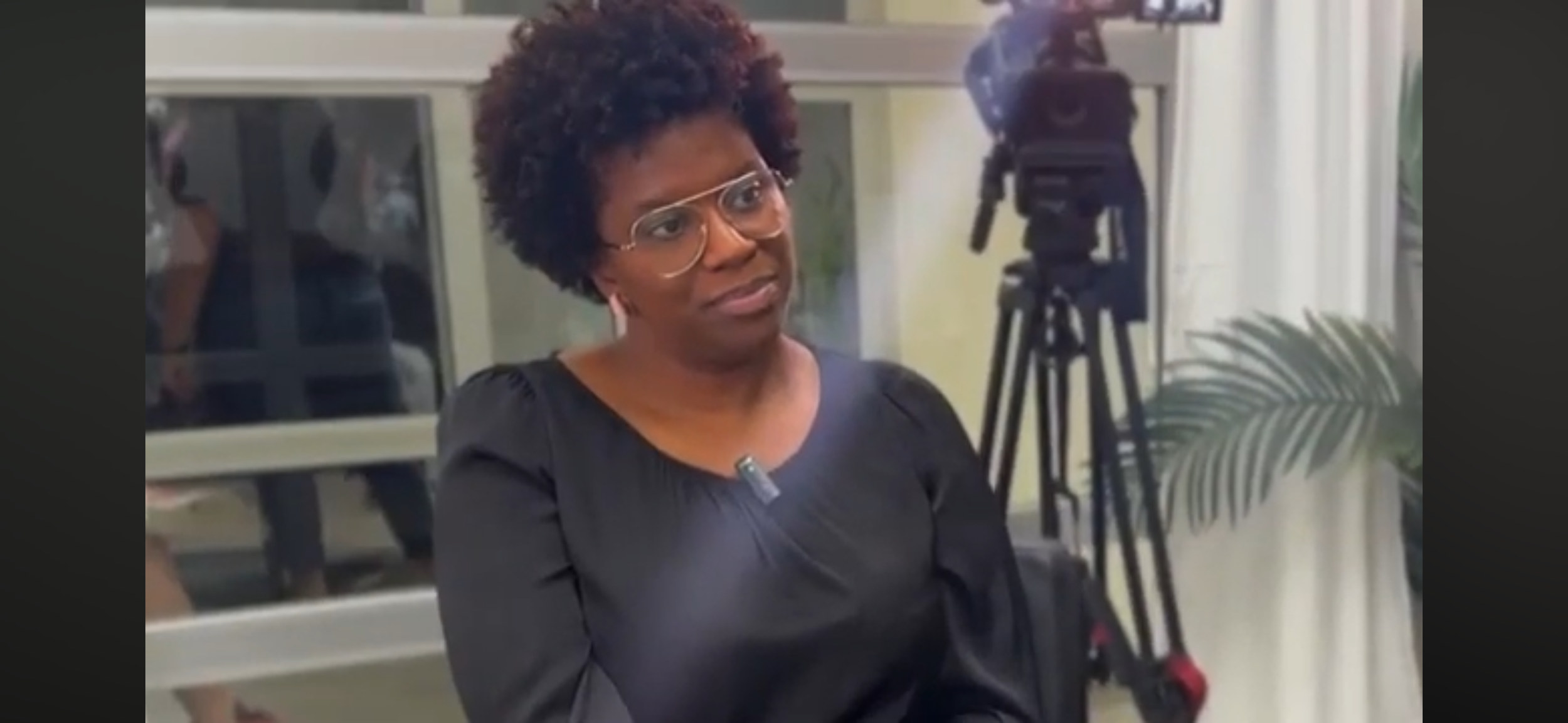 Latecia Hankerson wearing a black dress, appears to be speaking or listening during an interview or an event. A camera on a tripod and a person are visible in the background.