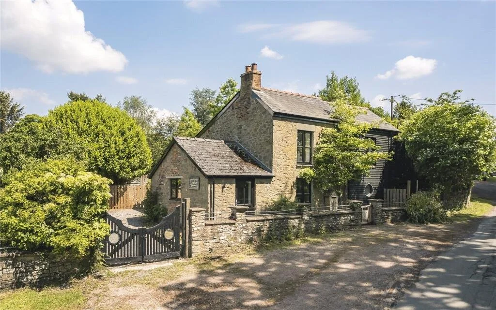 An old stone house with a gabled roof, surrounded by trees and a black fence with a gate, under a partly cloudy sky.