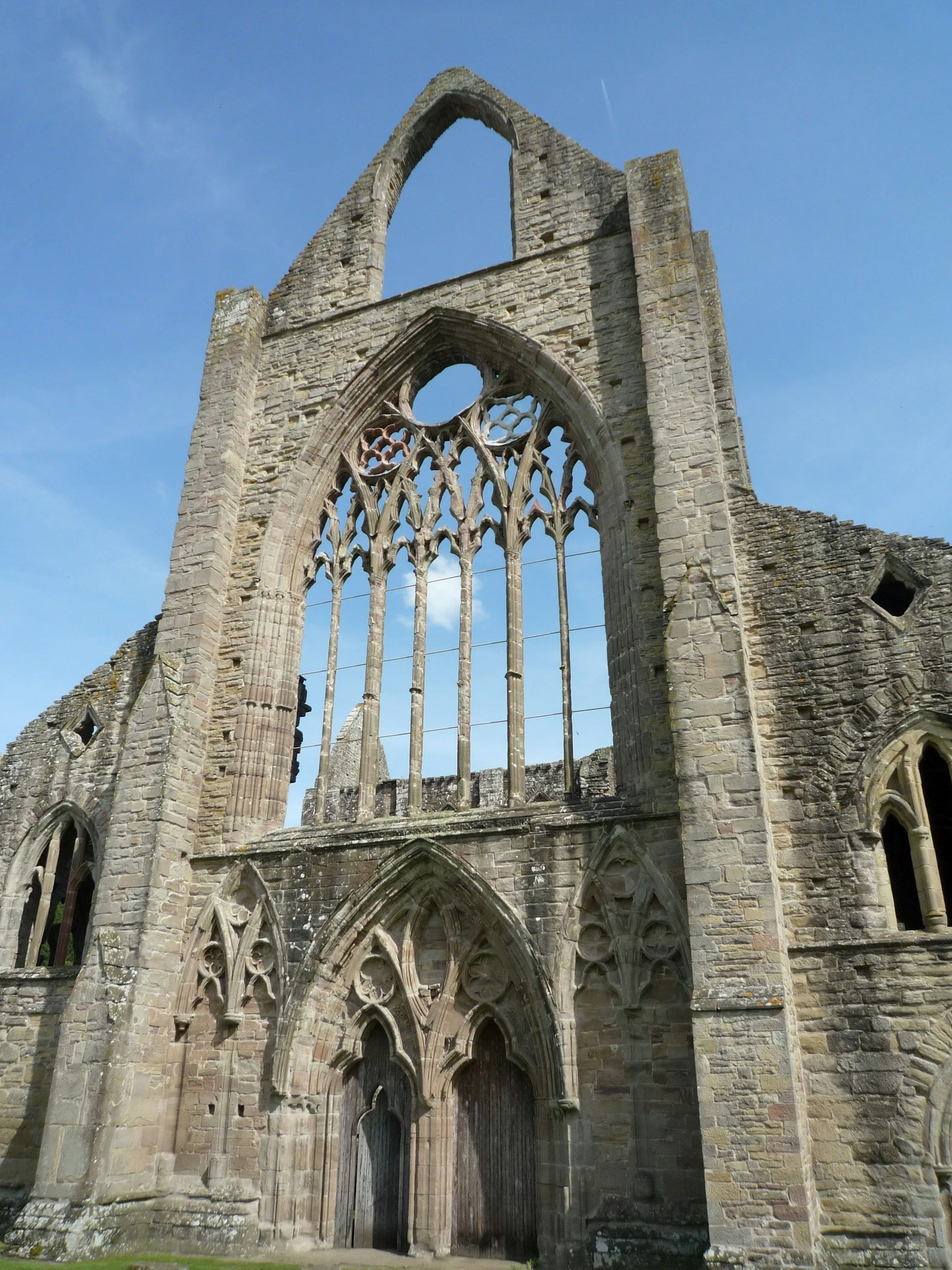 Ruins of a medieval stone church with large arched windows and an ornate doorway, set against a bright blue sky.