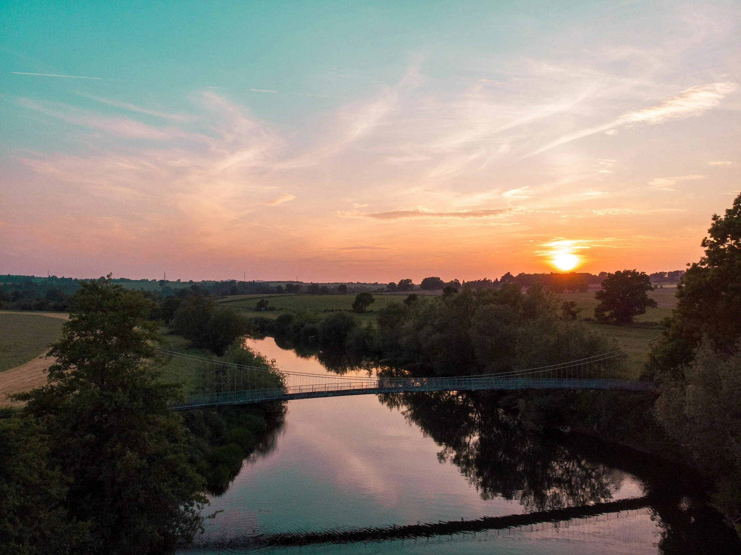 Sunset over a calm river with trees along the bank and a small suspension bridge in the foreground.
