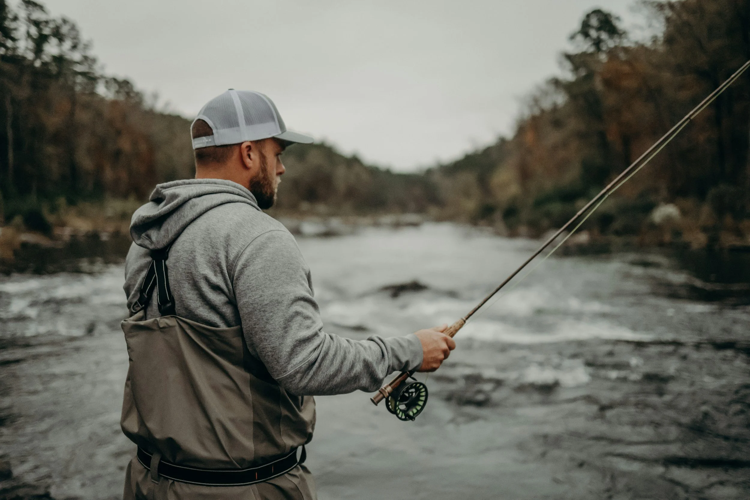 A man with a beard wearing a gray hoodie and a gray and white trucker hat fishing by a river, holding a fishing rod with both hands.