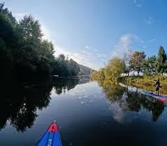 View of a calm river with trees on both sides, blue sky, and a person in a kayak on the right side.