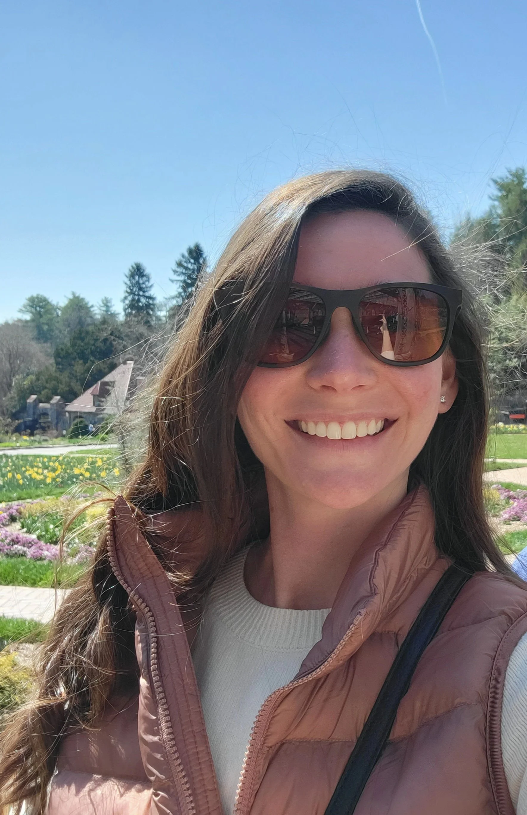 A woman smiling outdoors, wearing sunglasses, a beige sweater, and a brown quilted vest, with a park and houses in the background on a sunny day.