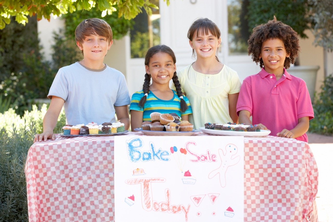 Four children standing behind a table with baked goods at a bake sale, outdoors with greenery and a house in the background, smiling at the camera.