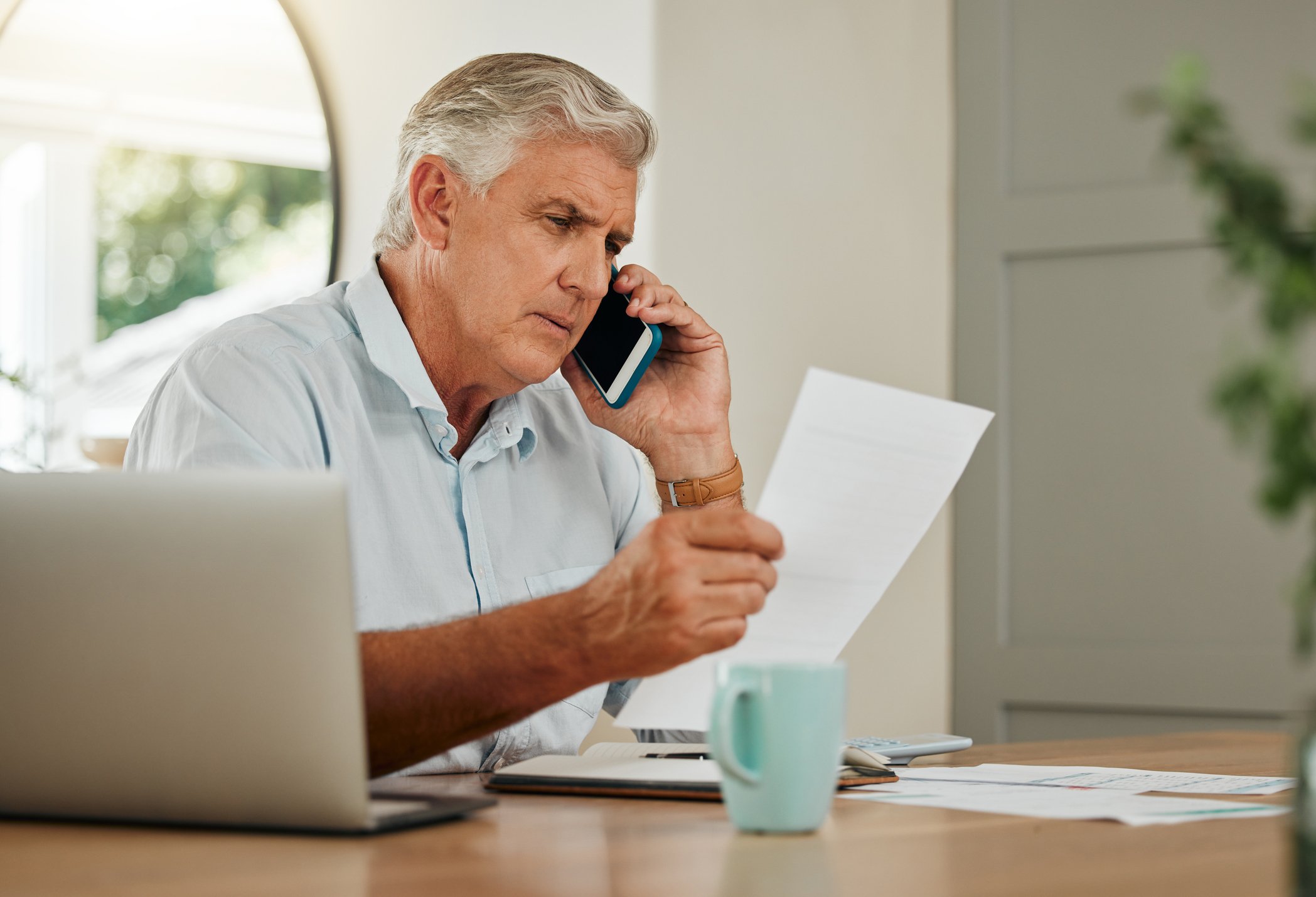 An elderly man with gray hair wearing a white shirt is sitting at a desk, holding a piece of paper in one hand and talking on his cell phone with the other. There is a white mug and some papers on the desk. The background shows a window with sunlight coming through.