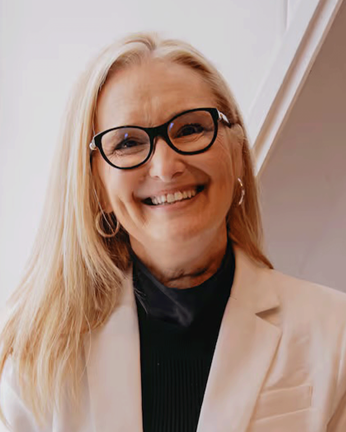 A smiling woman with blonde hair and glasses, wearing a cream blazer and black top, standing indoors.