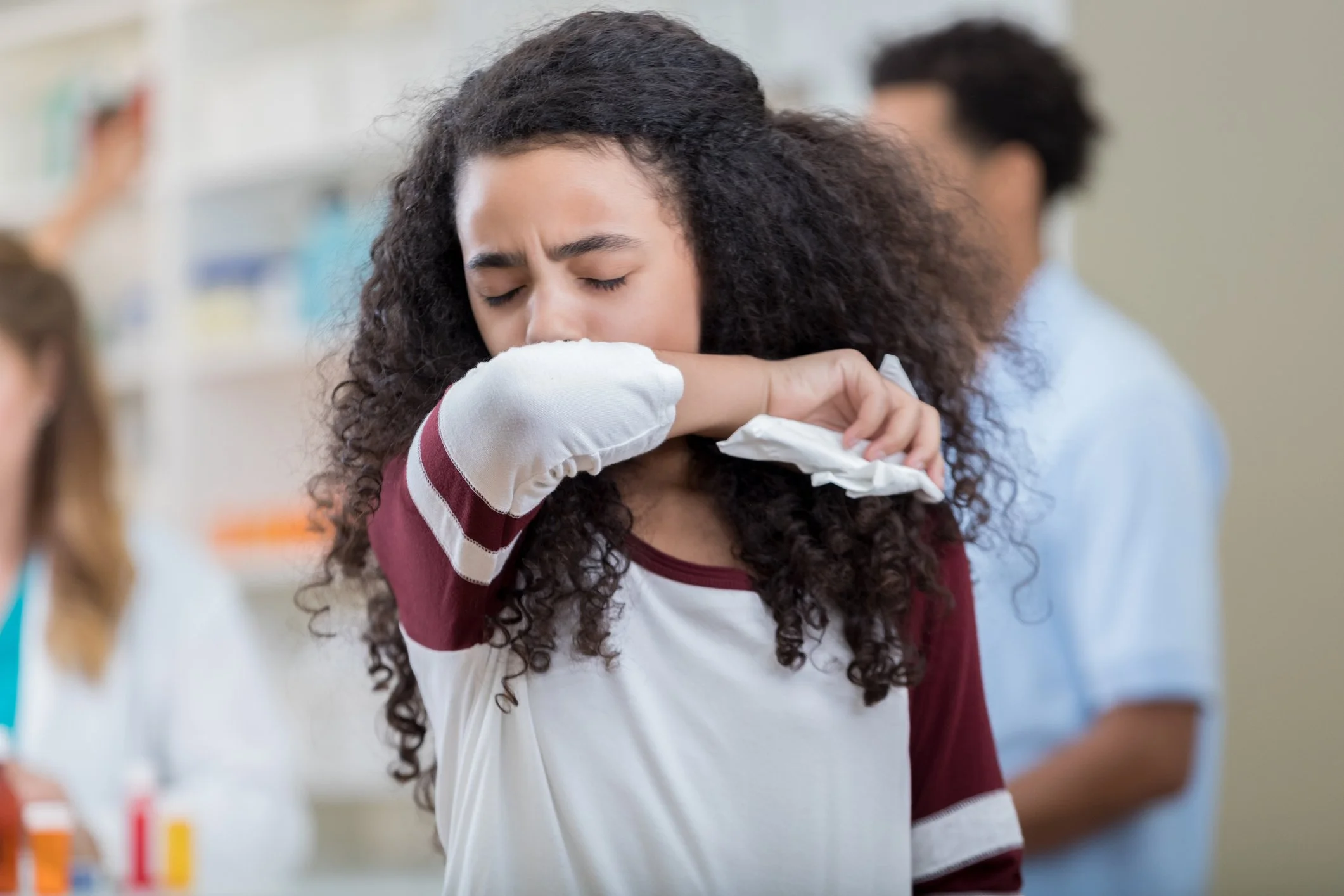 A young girl with curly hair is crying and hugging an arm with a tissue in a school setting.