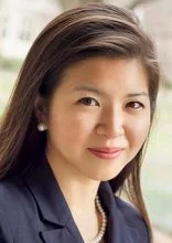 Close-up of a woman with long dark hair wearing a navy blazer and pearl earrings, smiling slightly.