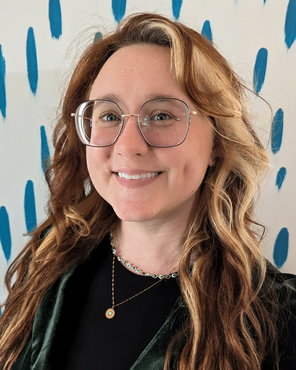A woman with wavy red hair, glasses, and a black shirt, smiling against a white wall with blue brushstroke patterns.