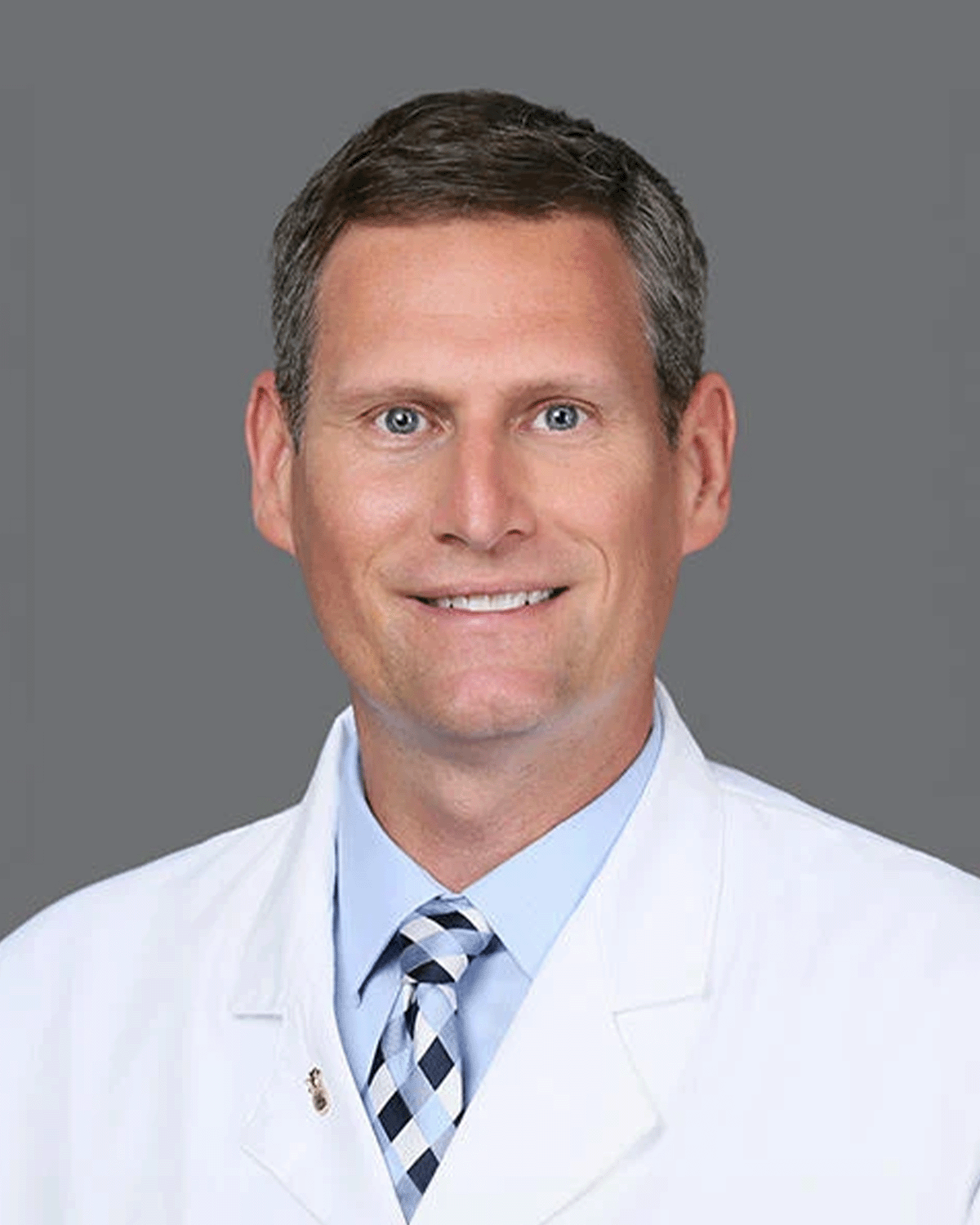 Headshot of a smiling male doctor with short brown hair, blue eyes, wearing a white coat, light blue shirt, and patterned tie, against a gray background.