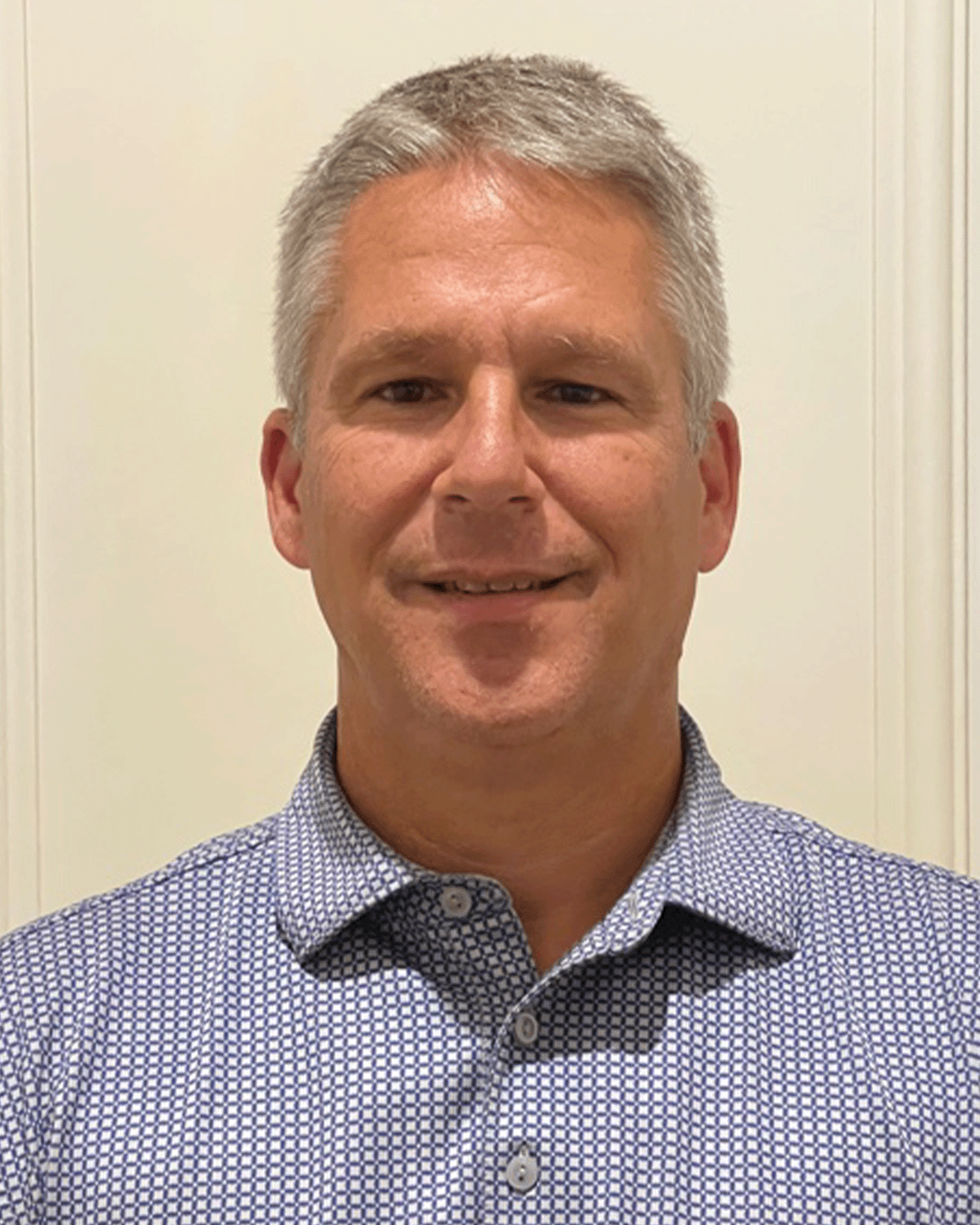 A middle-aged man with gray hair smiling, wearing a blue and white checkered shirt, standing in front of a light-colored wall.