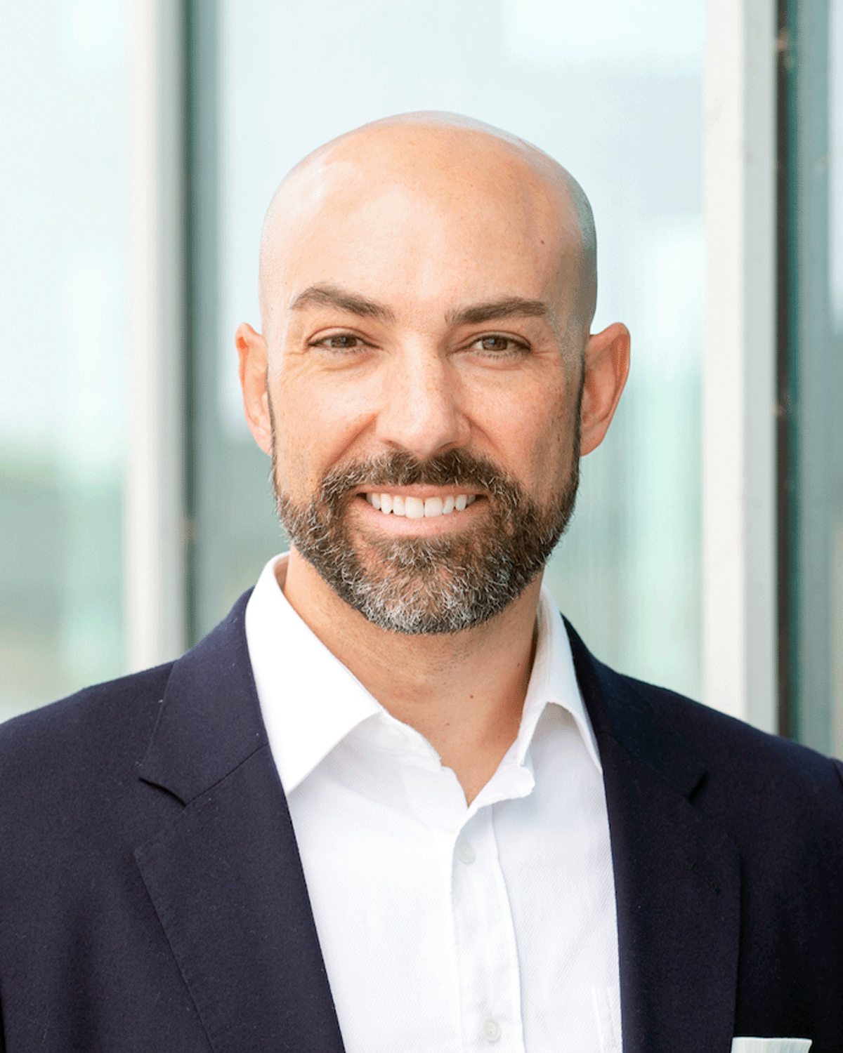 Headshot of a smiling man with a beard and a shaved head, wearing a dark suit jacket and white shirt, standing in front of a glass window.