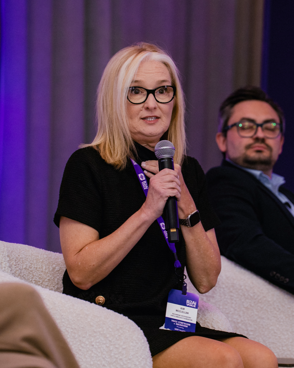 A woman with blonde hair, glasses, in a black dress, sitting on a white couch, holding a microphone, speaking at a conference or panel.