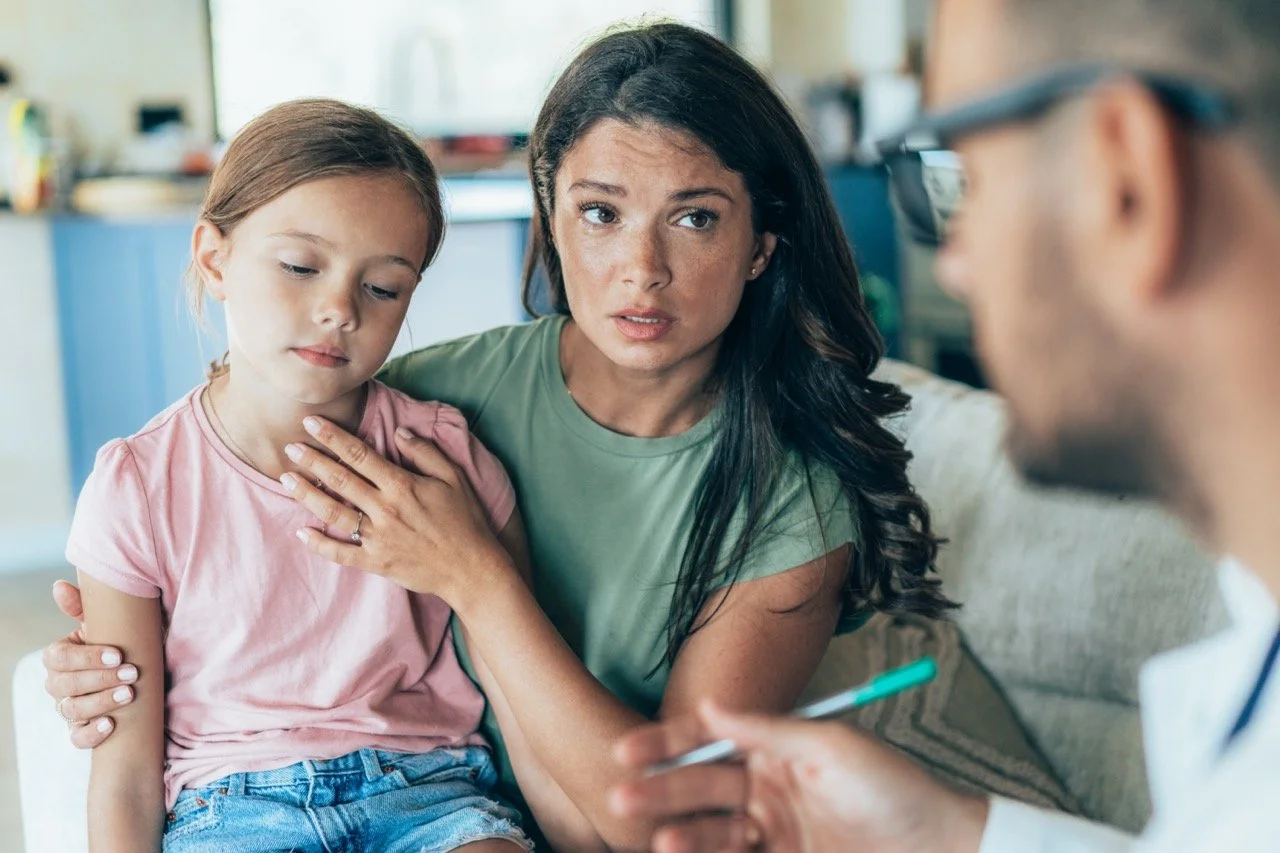 A young girl with a sad expression sitting on her mother's lap during a doctor's appointment, while a doctor with glasses speaks to them and holds a pen and notepad.