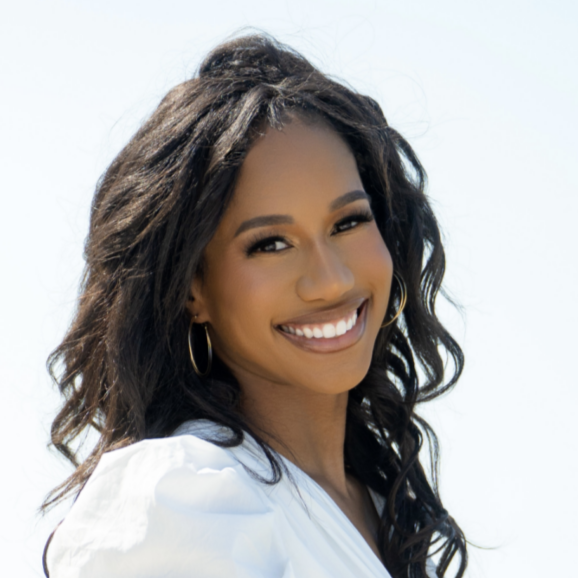 A smiling woman with curly dark hair and hoop earrings, wearing a white top against a clear sky background.