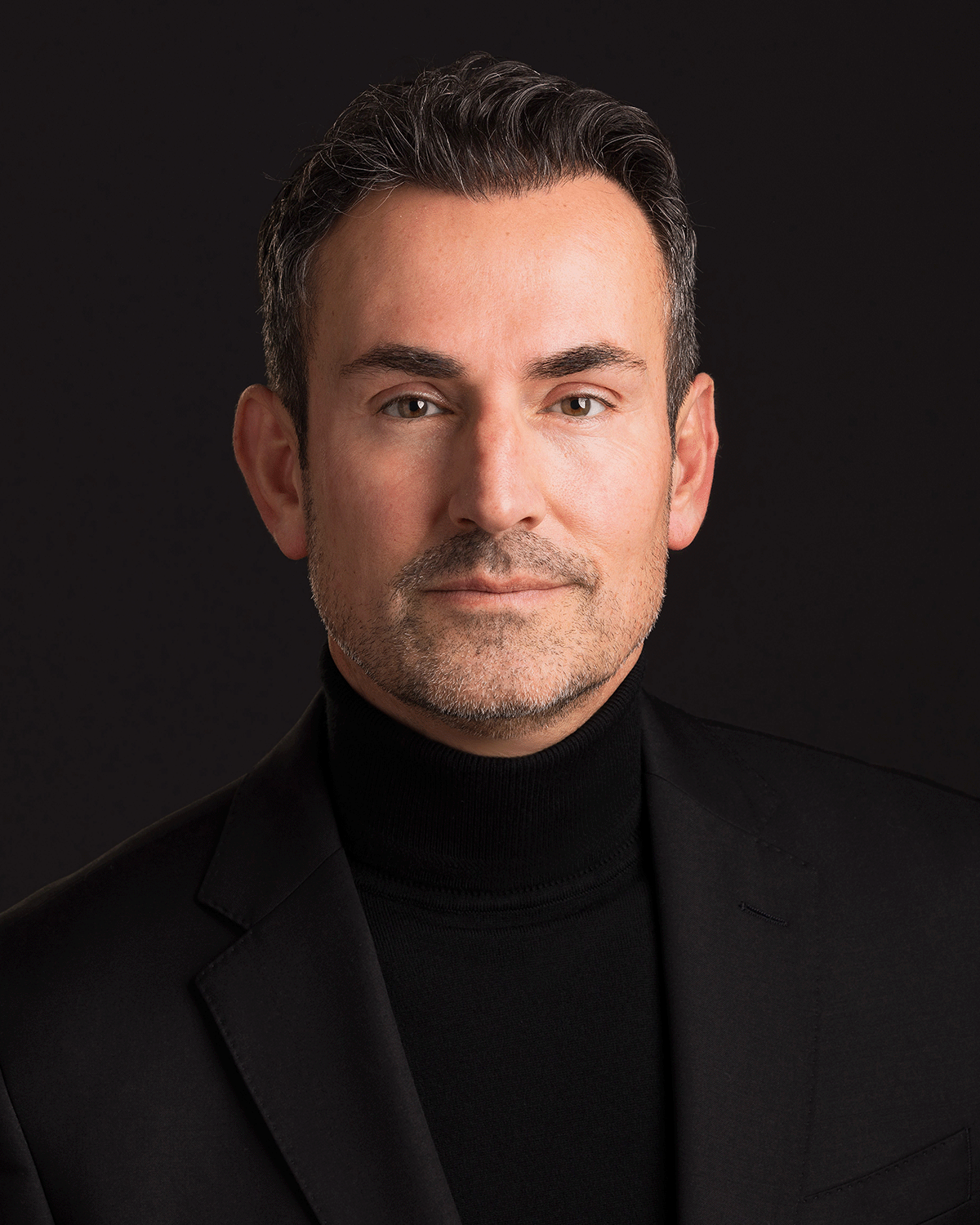 A professional man with short dark hair, light beard, wearing a black turtleneck and black blazer, posing against a black background.