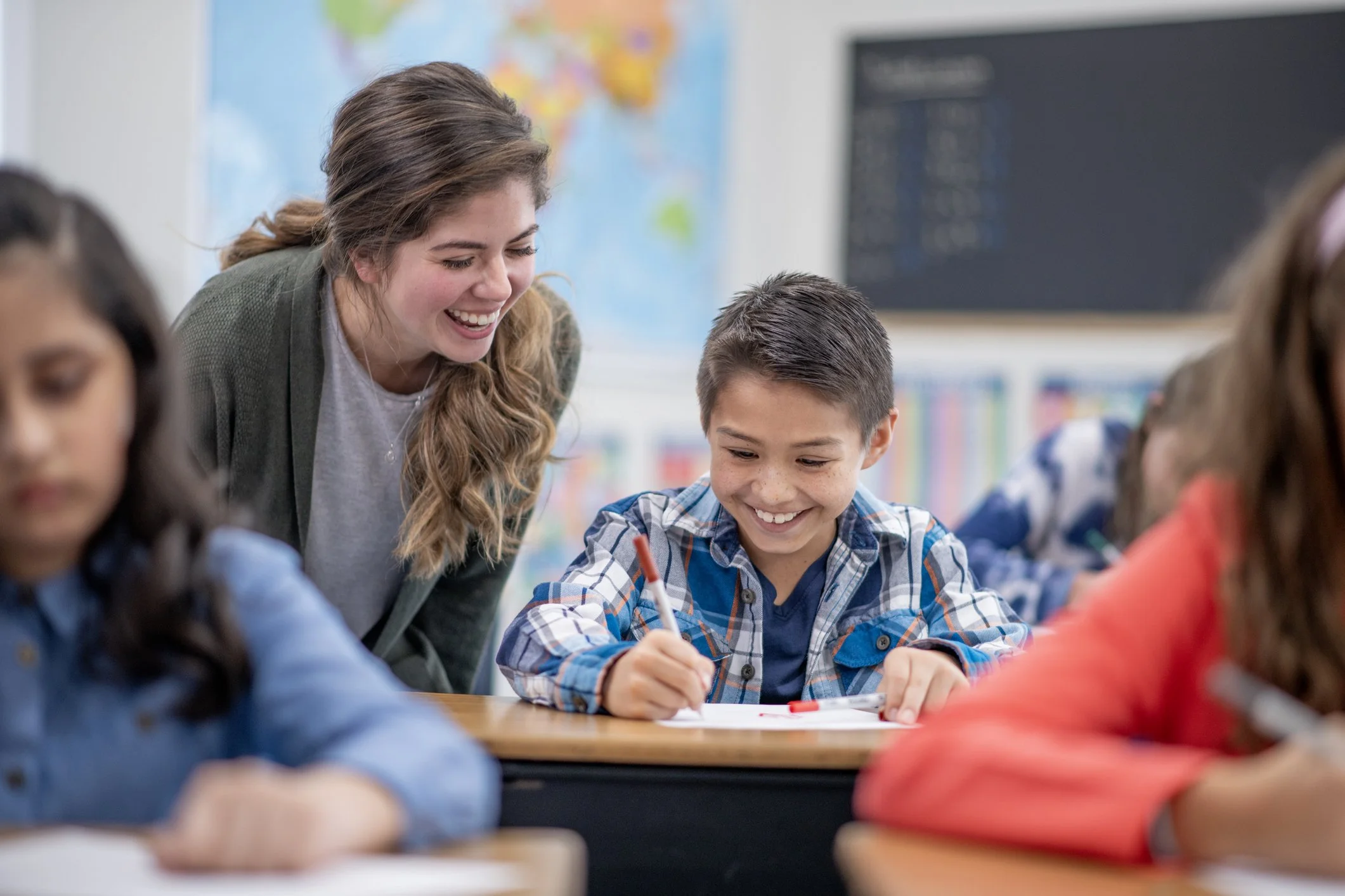 A classroom scene with a smiling female teacher leaning over to assist a male student in a plaid shirt, with other students blurred in the foreground and a world map on the wall.