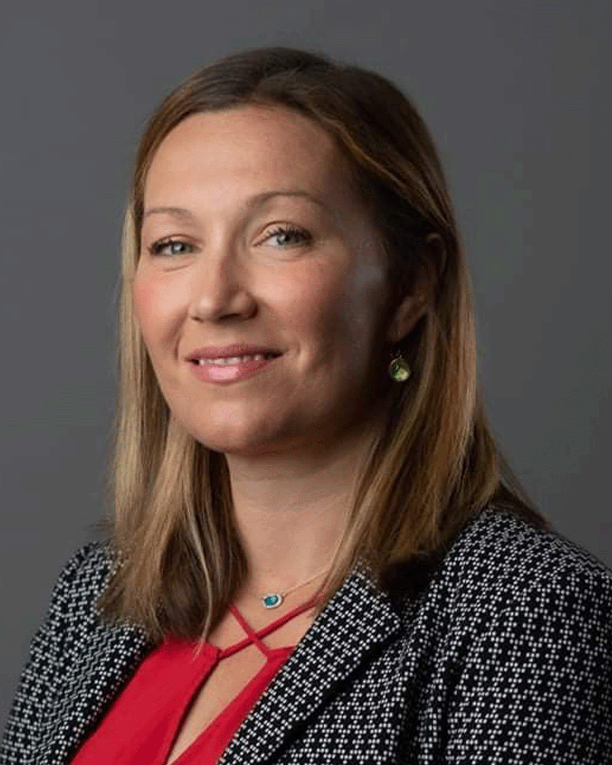 A professional woman with shoulder-length light brown hair, wearing a black and white patterned blazer over a red top, earrings, and a necklace, smiling against a gray background.