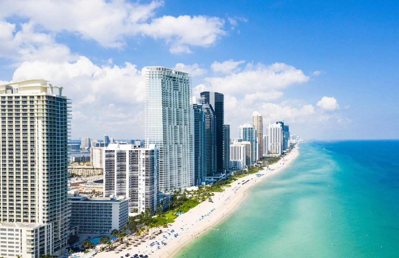 Aerial view of a sunny beach with high-rise buildings lining the coast and clear turquoise ocean waters.