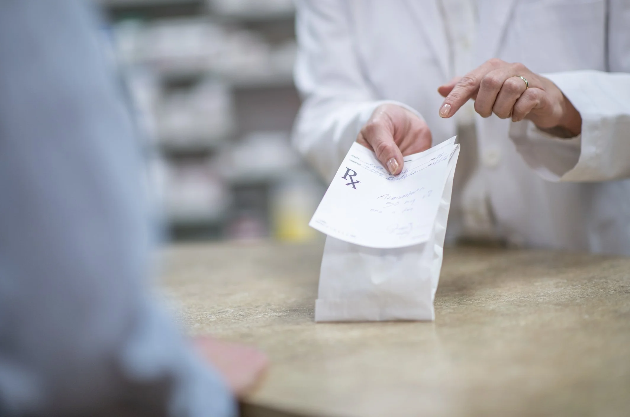 Person in a white coat holding a prescription prescription paper at a pharmacy counter.
