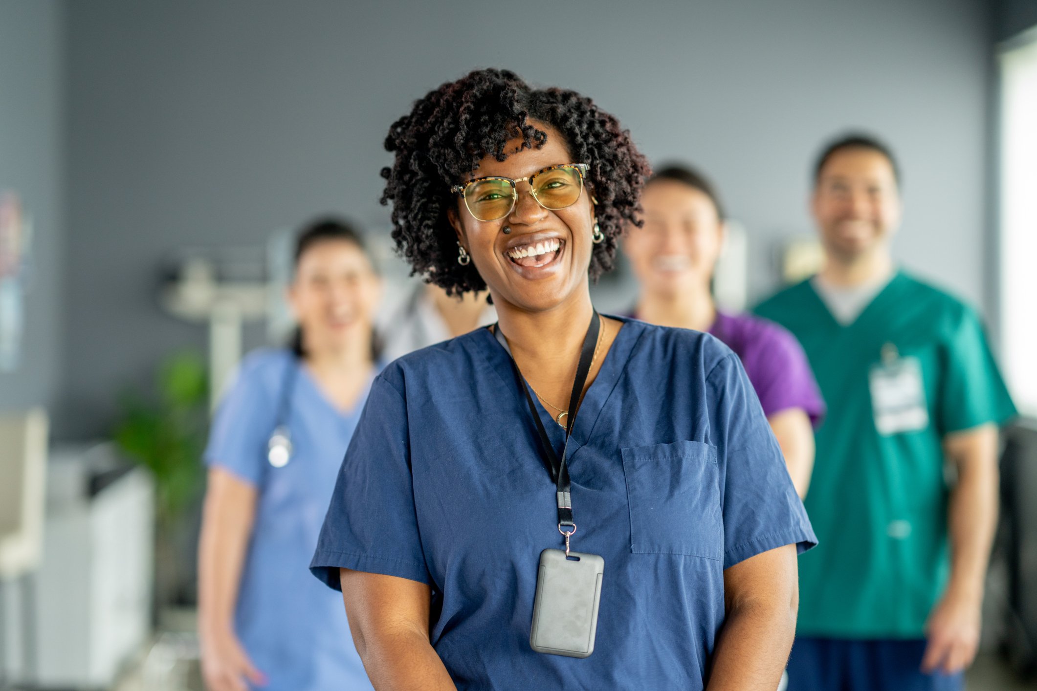 A group of healthcare workers, including a smiling woman with curly hair and glasses in the foreground, wearing scrubs and a stethoscope, in a medical setting.