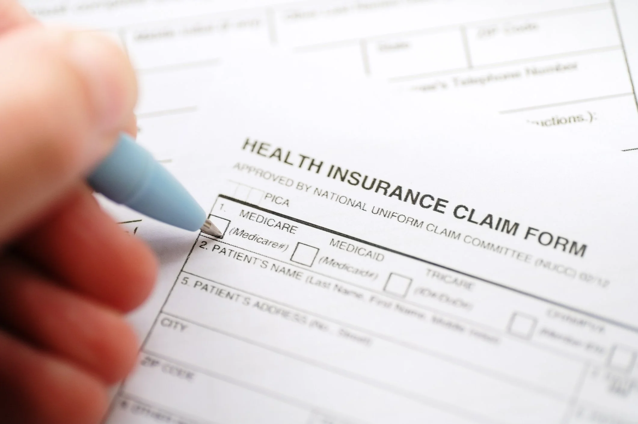 Close-up of a person filling out a health insurance claim form with a blue pen, focusing on the form titled 'HEALTH INSURANCE CLAIM FORM' with various sections for patient and insurance information.