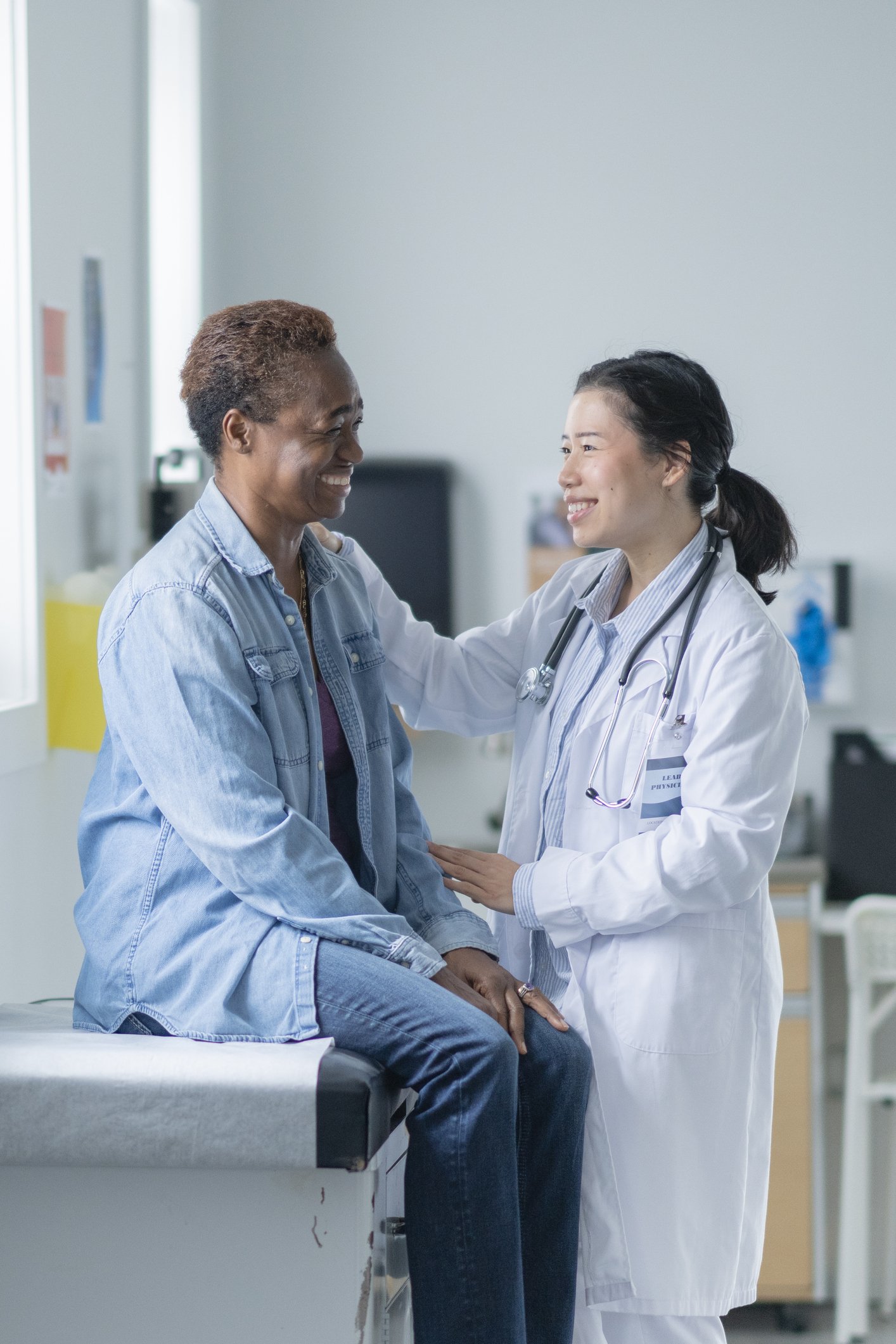 A female doctor in a white coat with a stethoscope around her neck comforting a woman sitting on an examination table in a medical office or clinic.