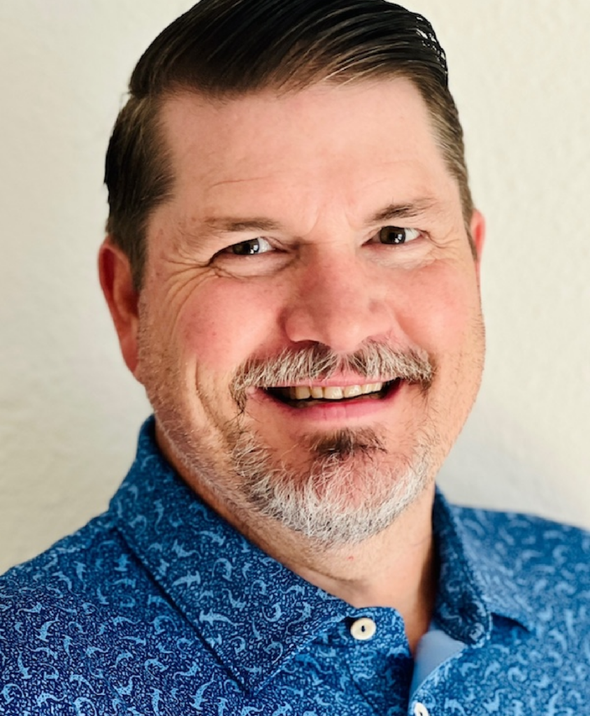 A smiling middle-aged man with a beard and mustache, wearing a blue patterned shirt, standing in front of a plain light-colored background.