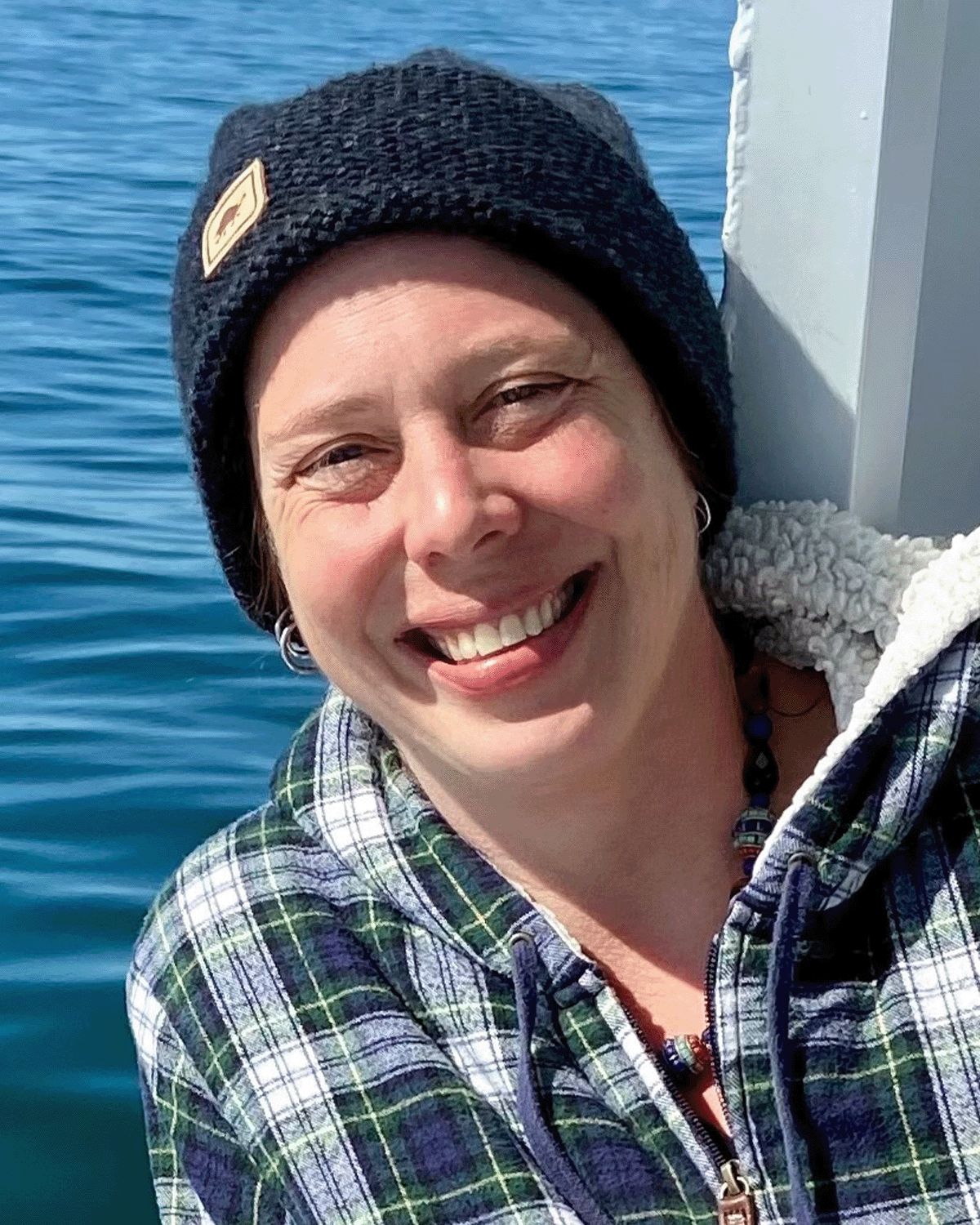 A smiling woman with short hair wearing a black knit hat, earrings, a necklace, and a plaid shirt, outdoors near water.