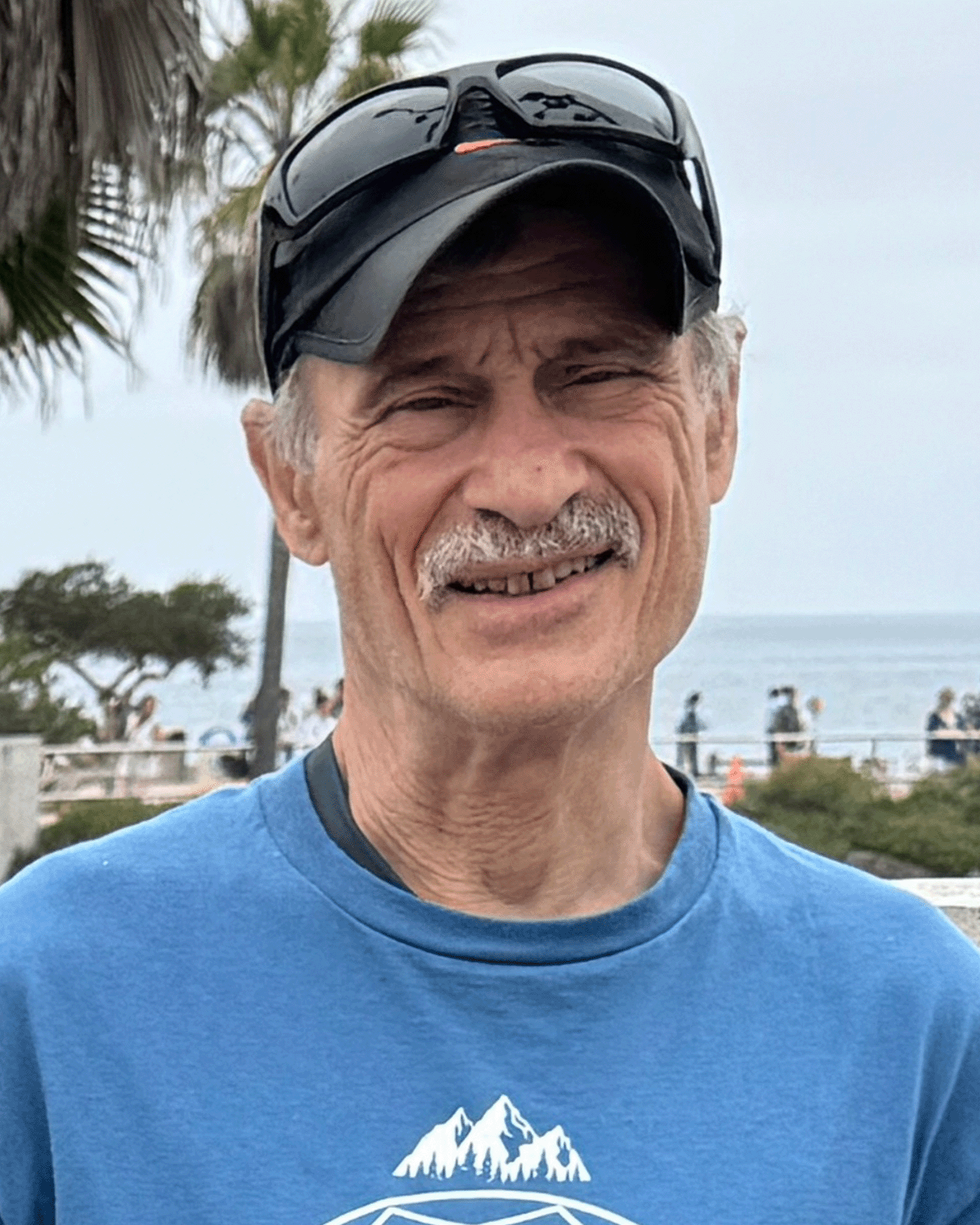 A smiling man wearing a baseball cap with sunglasses on top, in a blue t-shirt, outdoors by the water with trees and people in the background.
