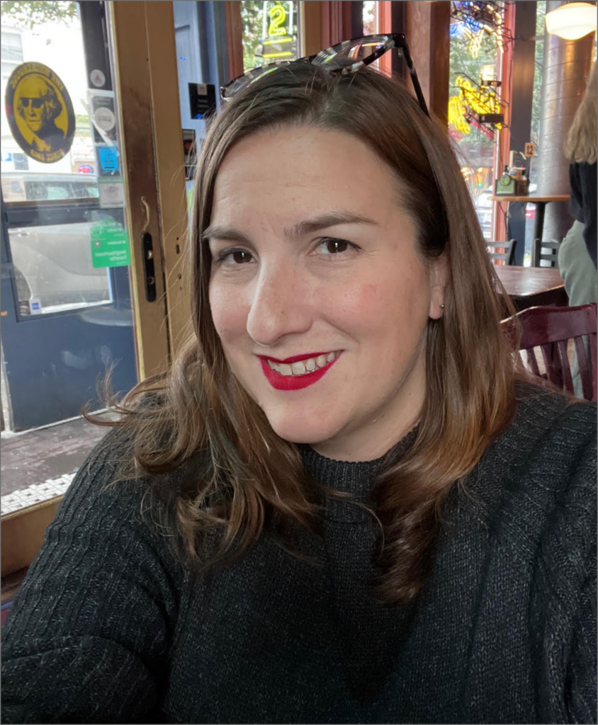 A woman with shoulder-length brown hair, wearing a black sweater and red lipstick, smiling at the camera inside a restaurant or cafe.